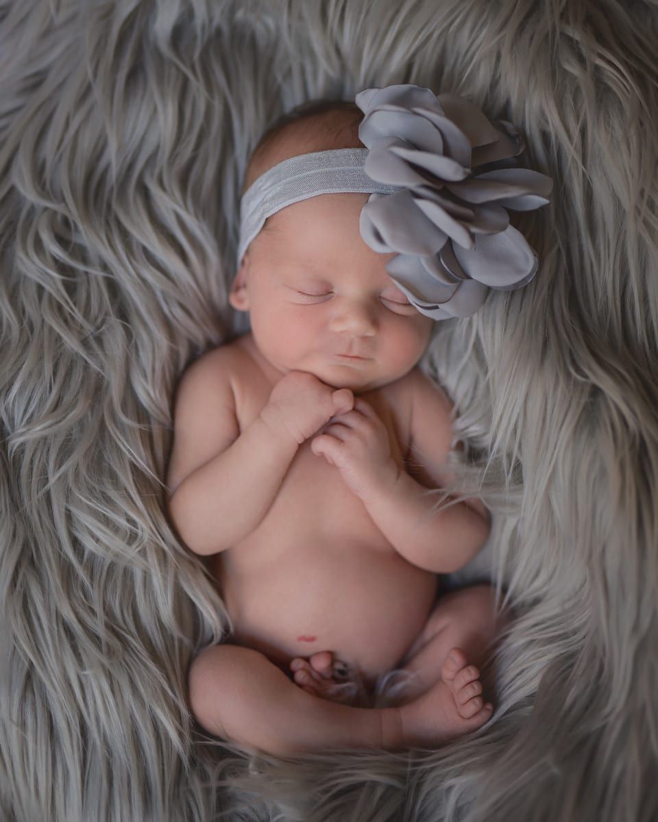 Newborn baby girl sleeping peacefully on a grey faux fur blanket; wearing grey headband with flower.