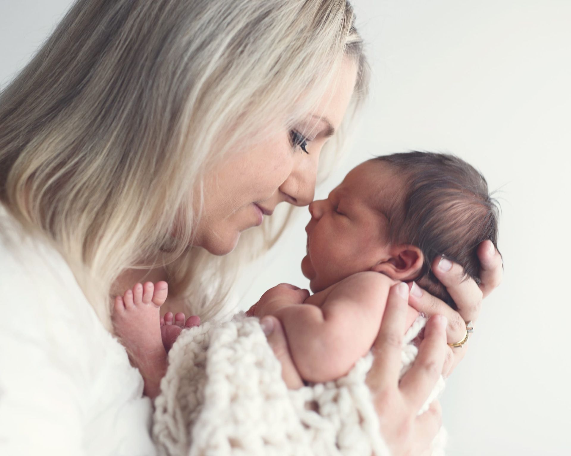 Mother with blonde hair gently holding a newborn baby close, both touching noses, against a white background.