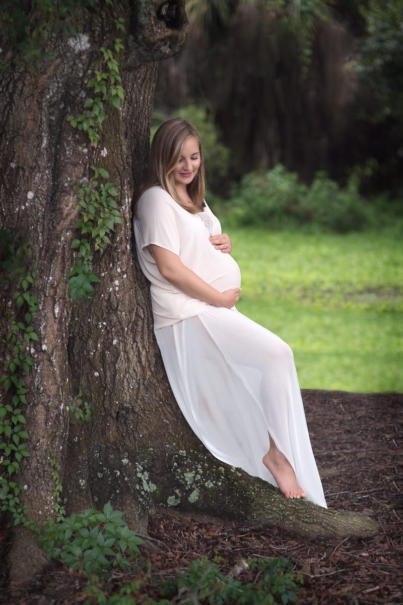Pregnant woman in white dress leaning against a tree, touching her belly outdoors.