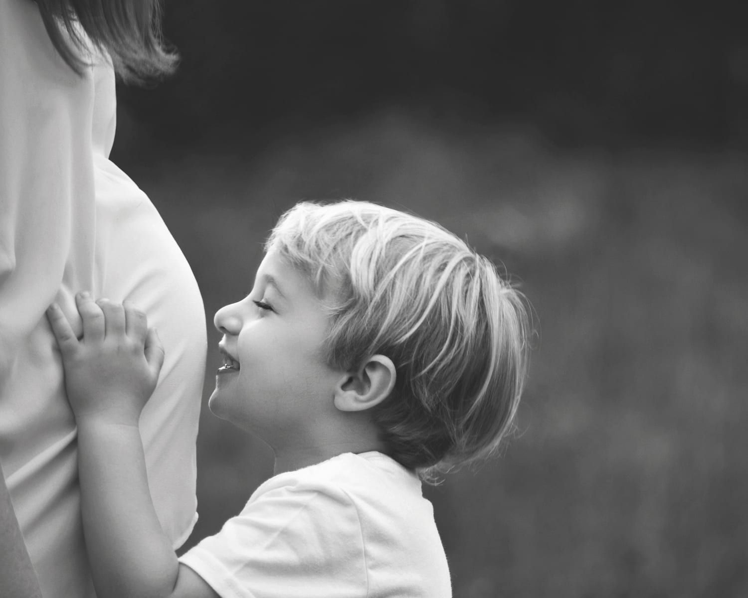 Blond child smiles, nuzzles pregnant belly in grayscale outdoors.