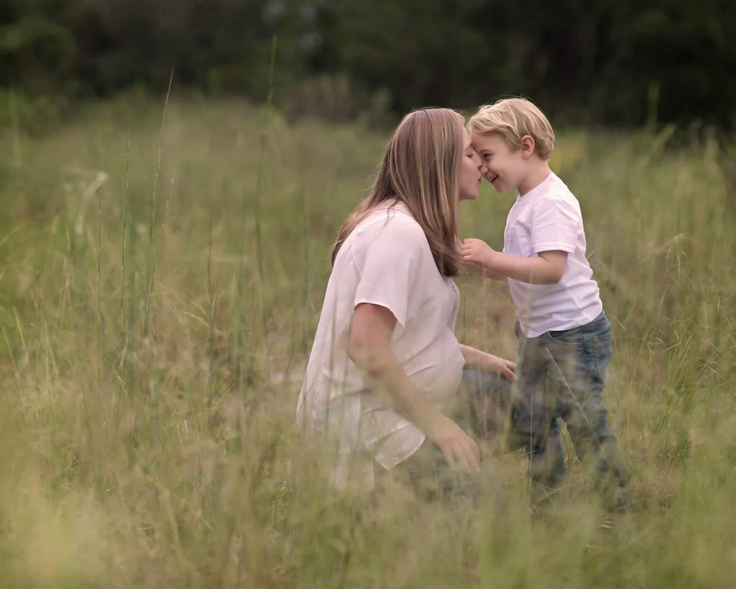 Pregnant woman kneels in field, kissing cheek of blond child.