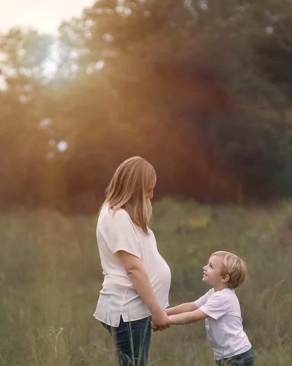 Pregnant woman holding hands with a young child in a field, backlit by the sun.