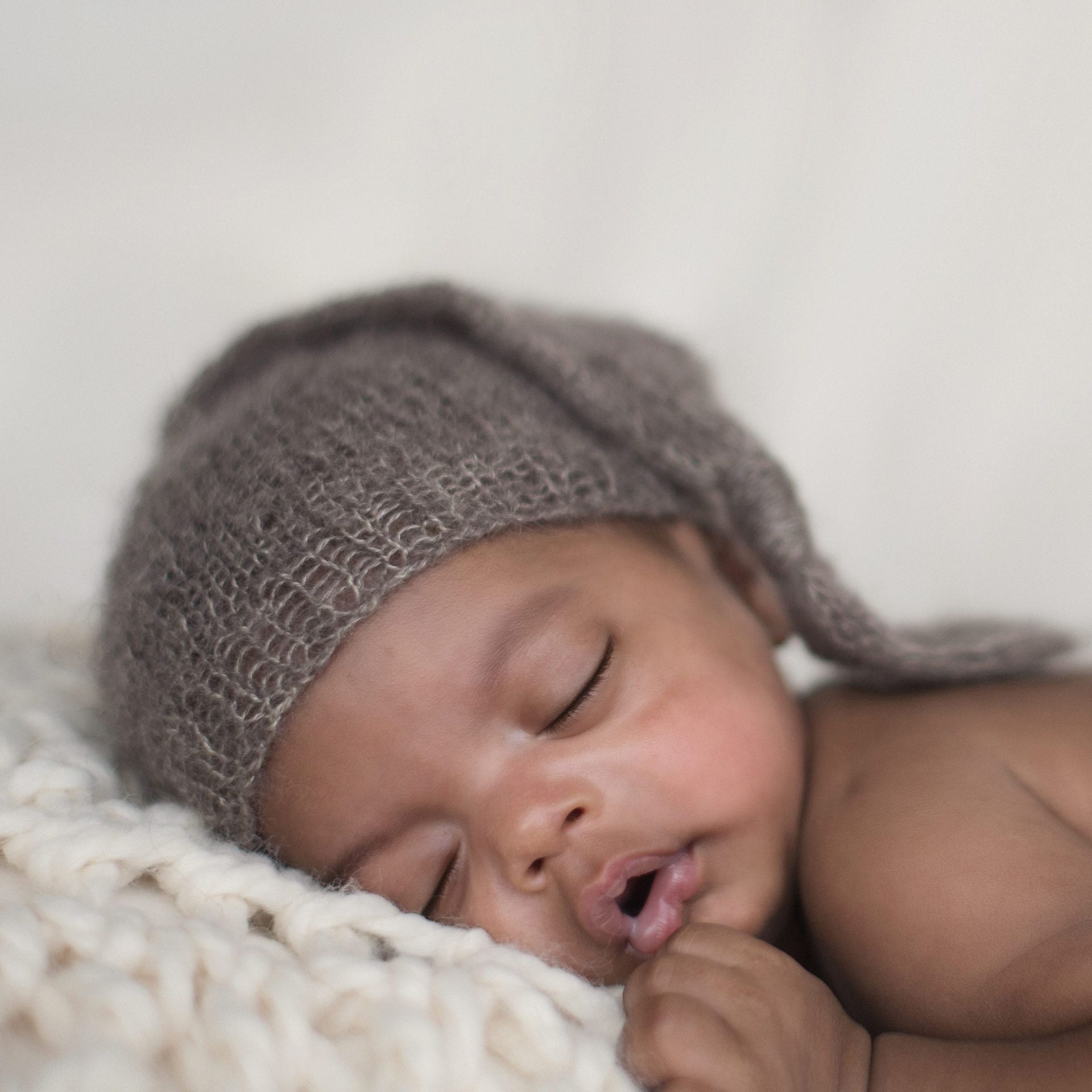 Sleeping baby wearing a gray knit cap, resting on a cream-colored blanket.