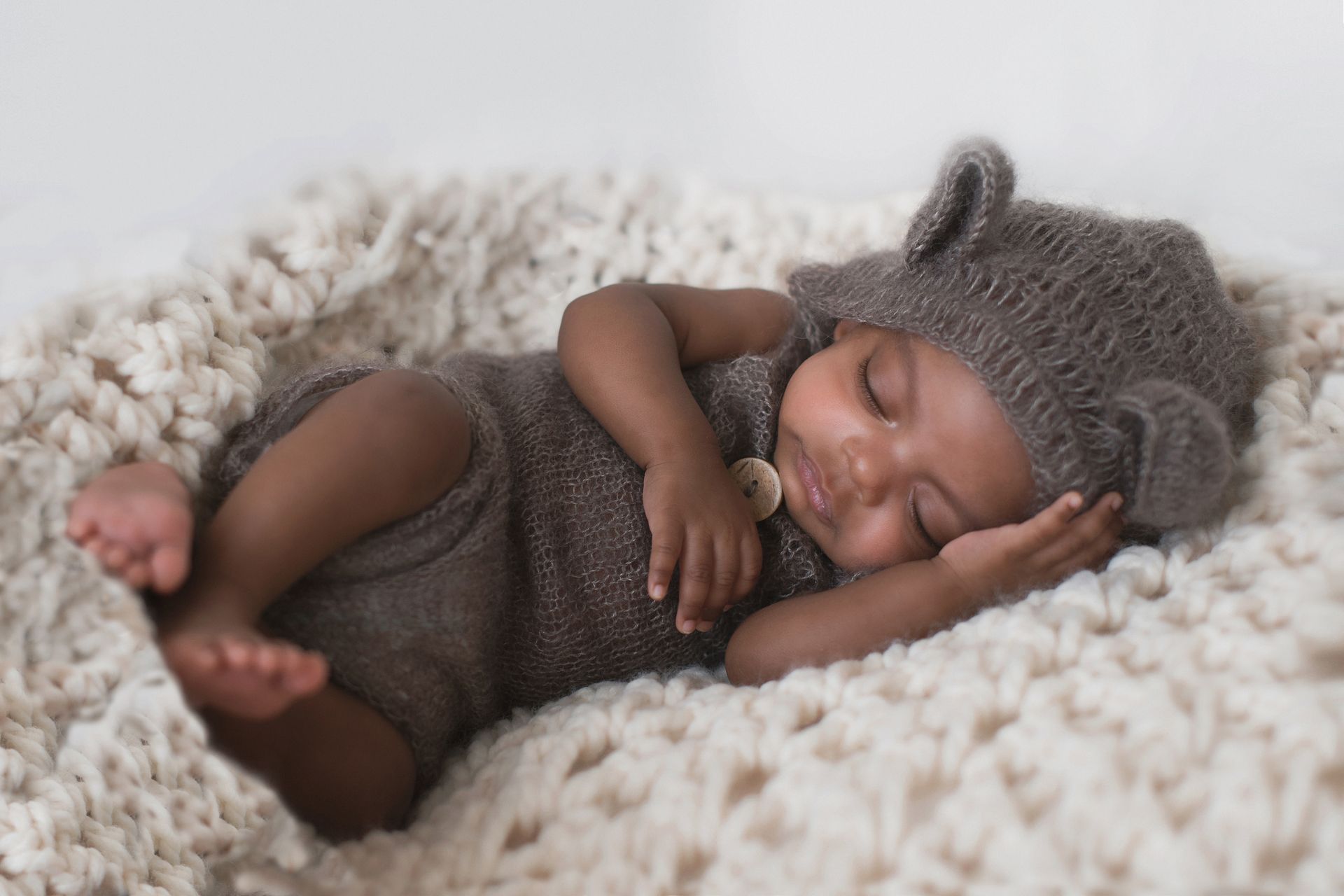 Sleeping baby wearing a bear hat and romper, nestled in a cream-colored blanket.