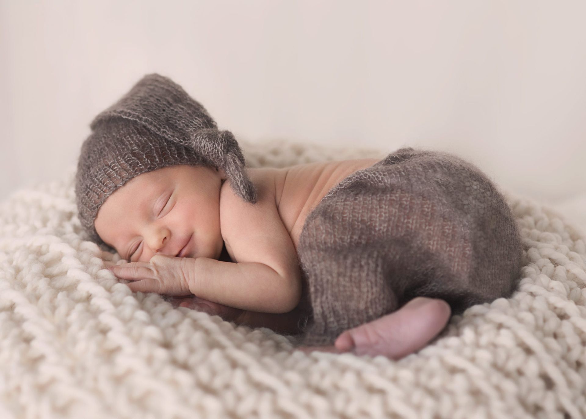 Newborn baby sleeping on a cream blanket, wearing brown knit cap and pants.