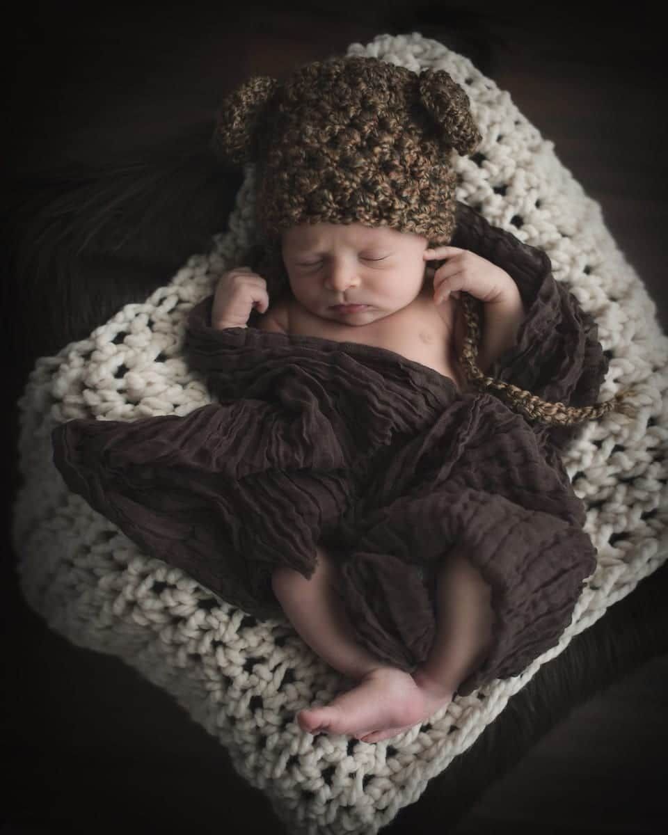 Newborn baby wearing a bear hat, swaddled in brown fabric, lying on a cream-colored blanket.