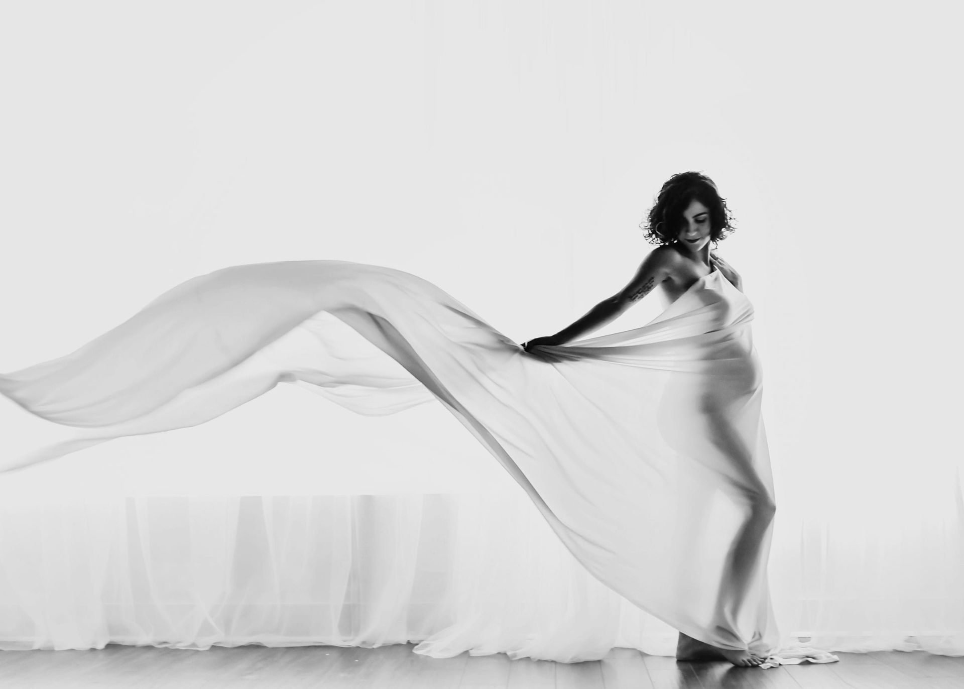Pregnant woman holding a flowing sheer fabric in a studio with bright lighting.