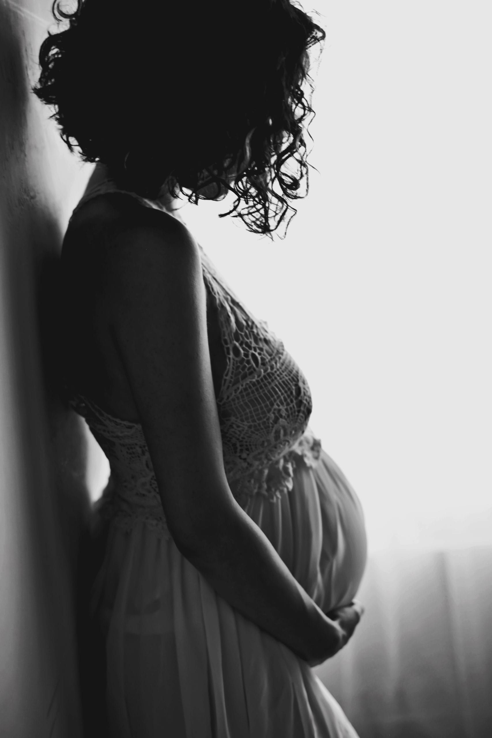 Pregnant woman in a flowing dress, leaning against a wall, hands cradling her belly.