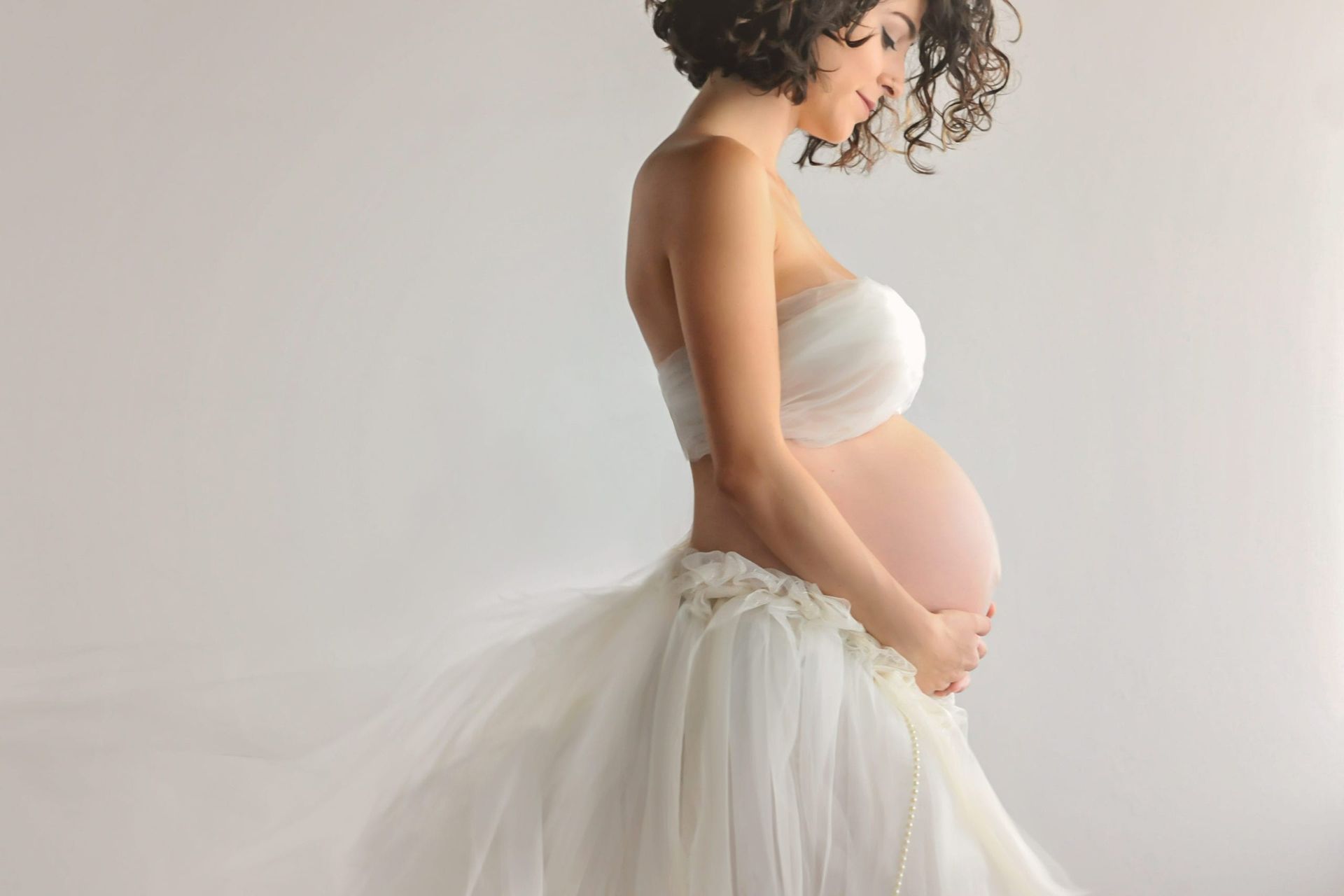 Pregnant woman in white strapless dress, embracing belly, side view, light background.