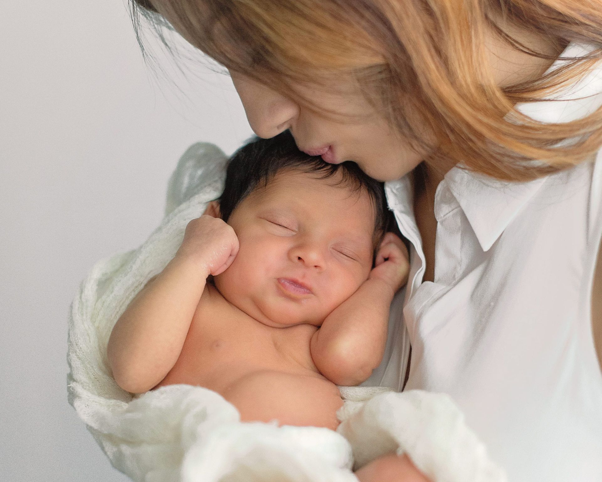 Woman kissing sleeping newborn wrapped in white blanket.