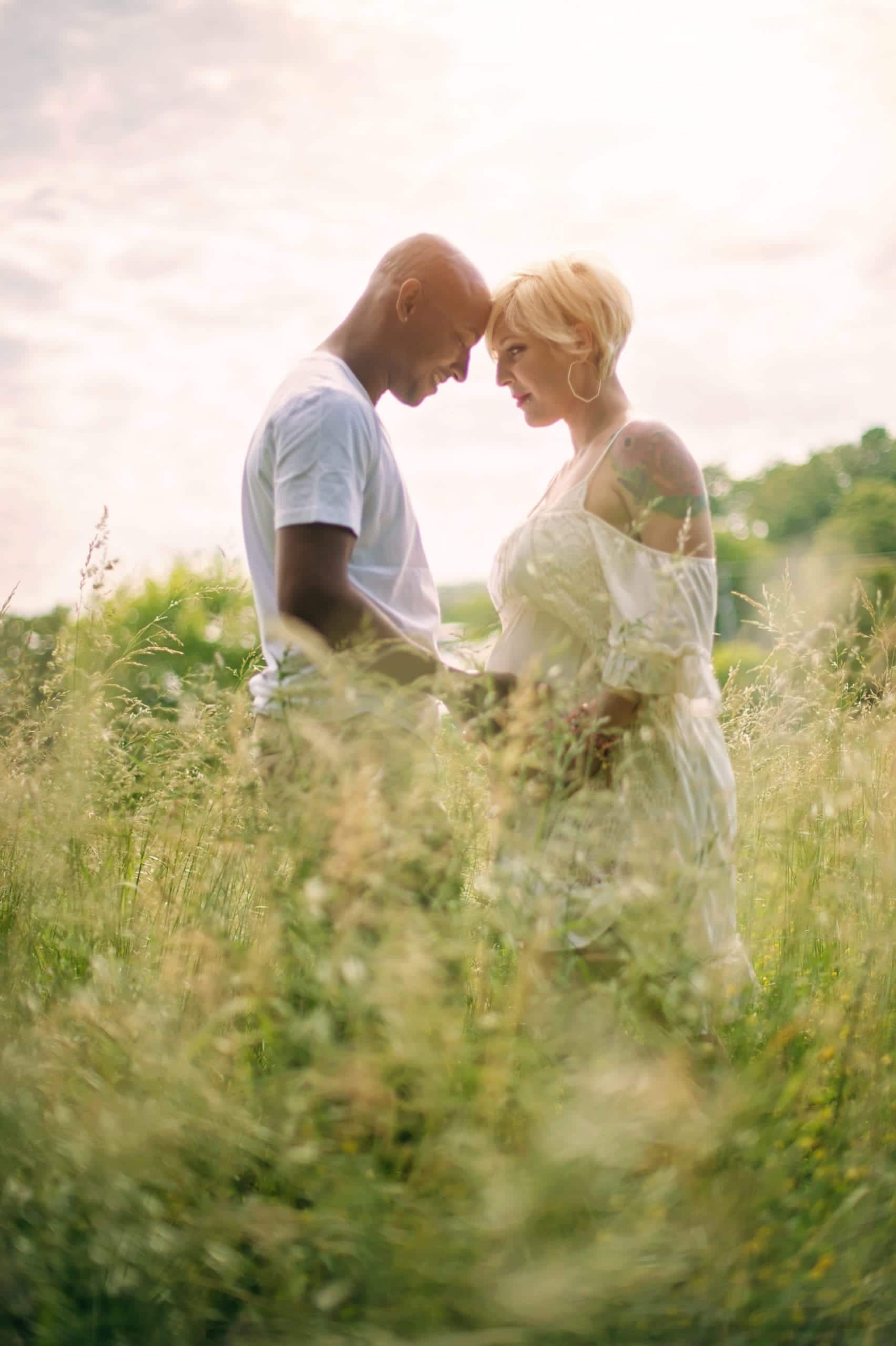 A biracial couple in a field touching foreheads, soft sunlight, the woman is wearing a white dress, and the man is wearing a white t-shirt.