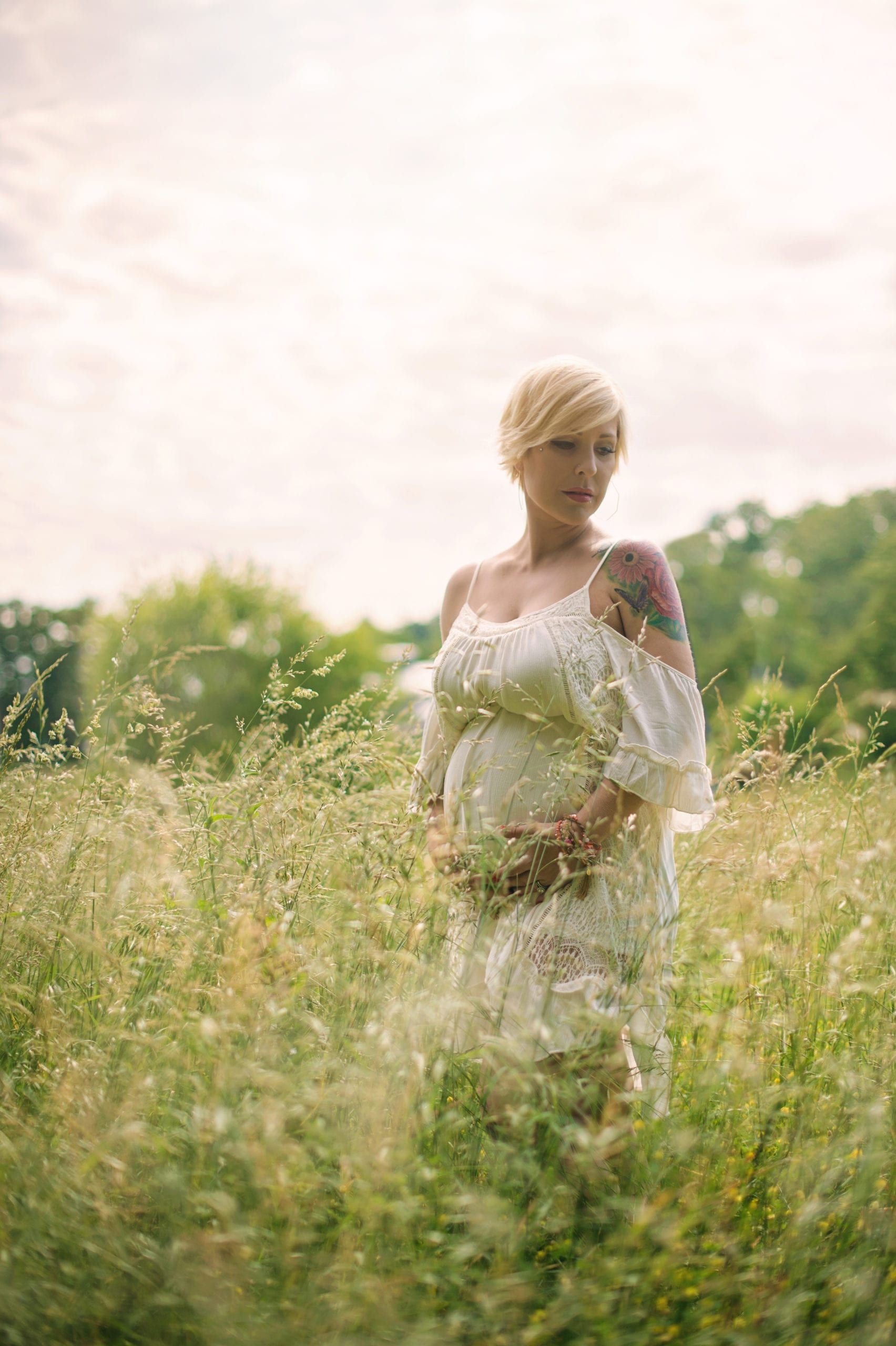 Pregnant woman in a white dress in a tall grassy field, holding her belly. Short blonde hair, tattoo on shoulder.
