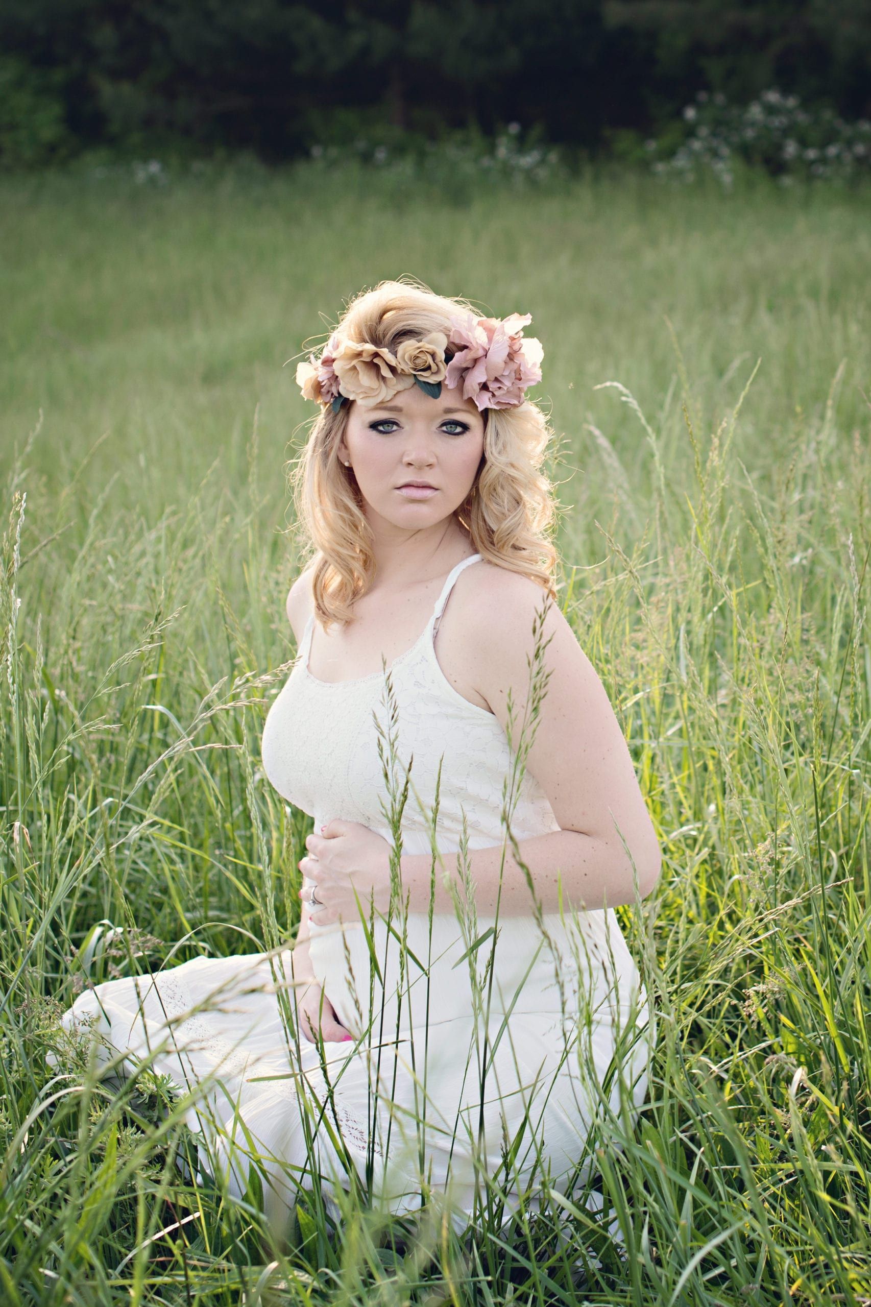 Blonde woman wearing flower crown and white dress, kneeling in tall grass, looking at the camera.
