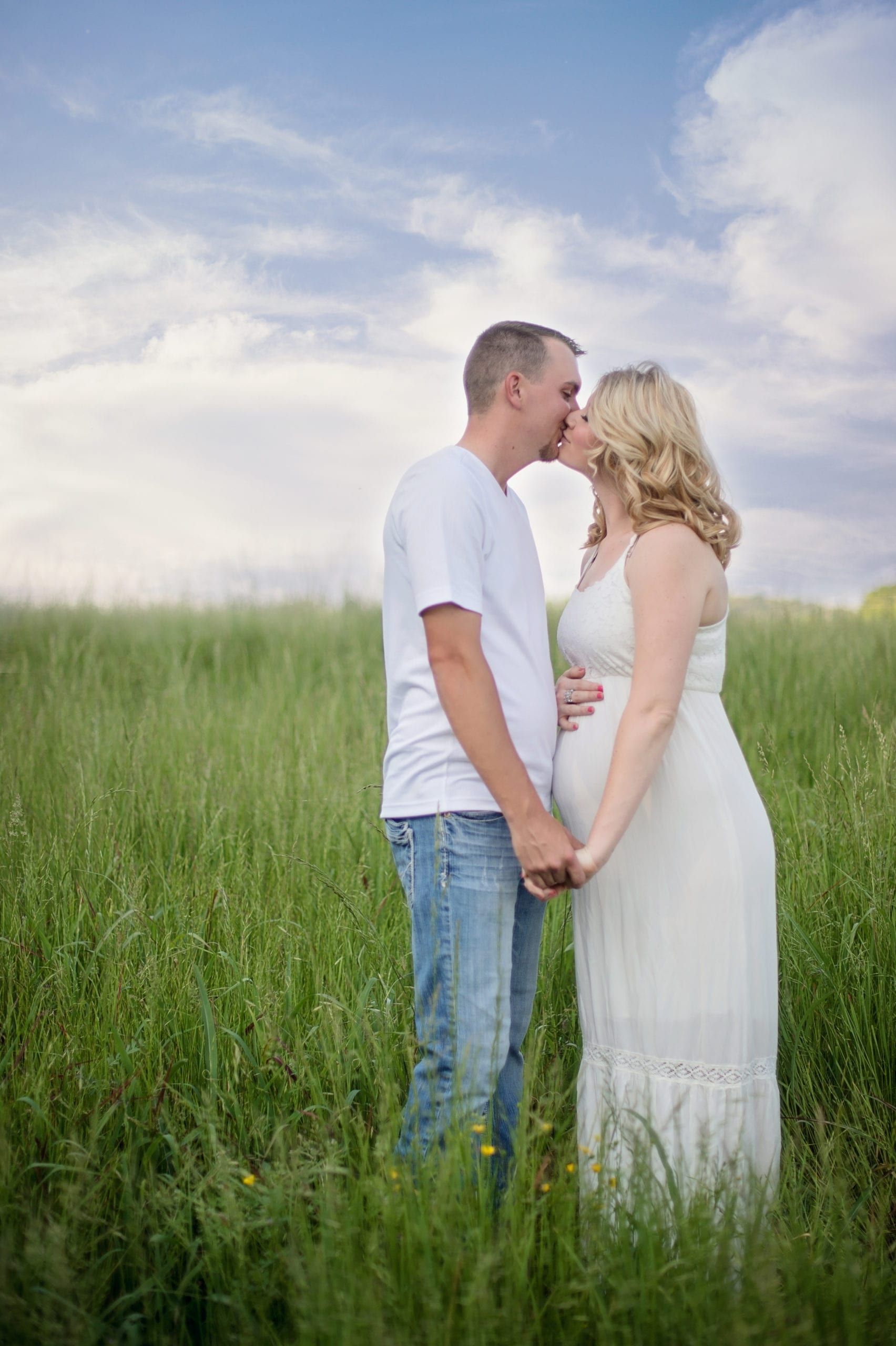Pregnant couple kissing in a tall green field under a cloudy sky.