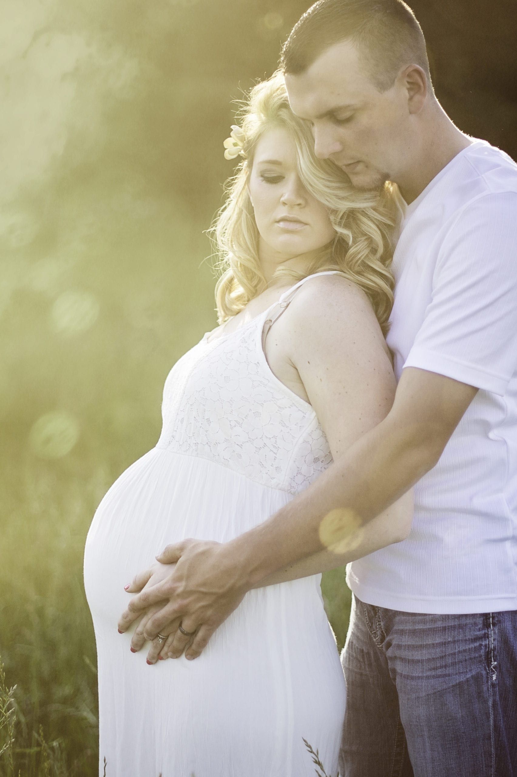 Pregnant woman in white dress and man embrace, touching her belly. Sunlight.
