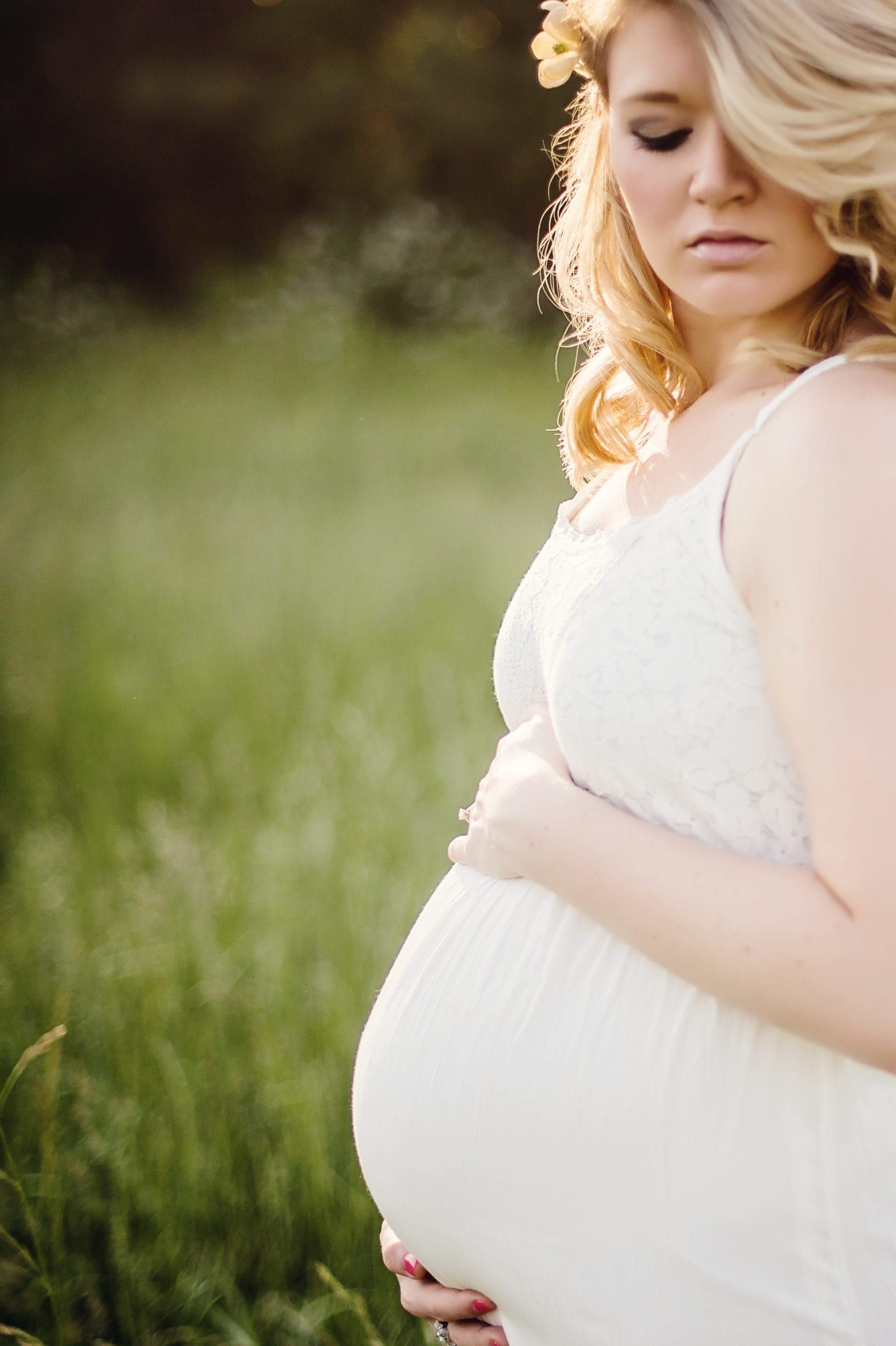 Pregnant woman in white dress, cradling her belly, standing in a grassy field, wearing flower crown.