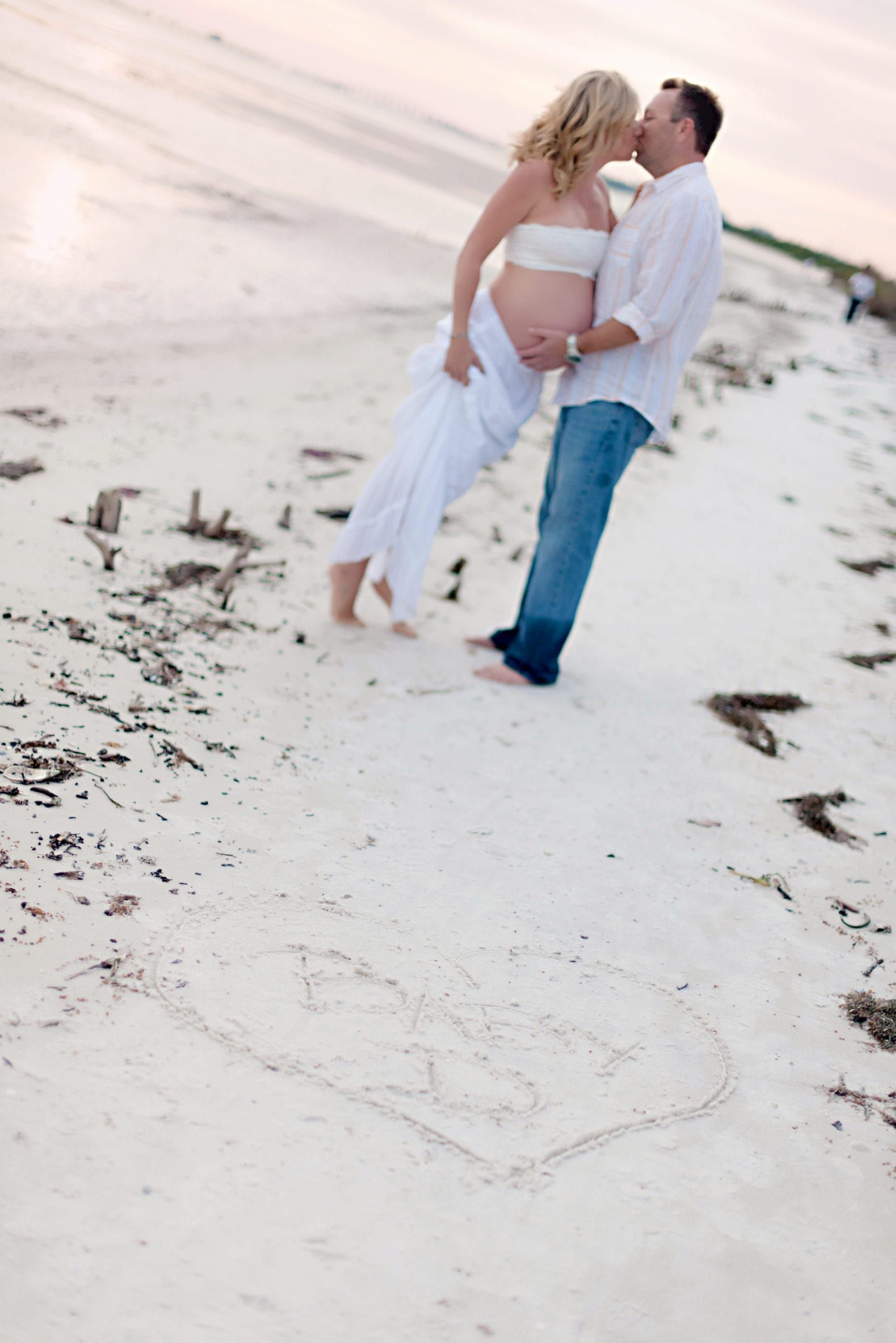 Pregnant woman and man kissing on a beach, holding baby bump, heart drawn in sand.
