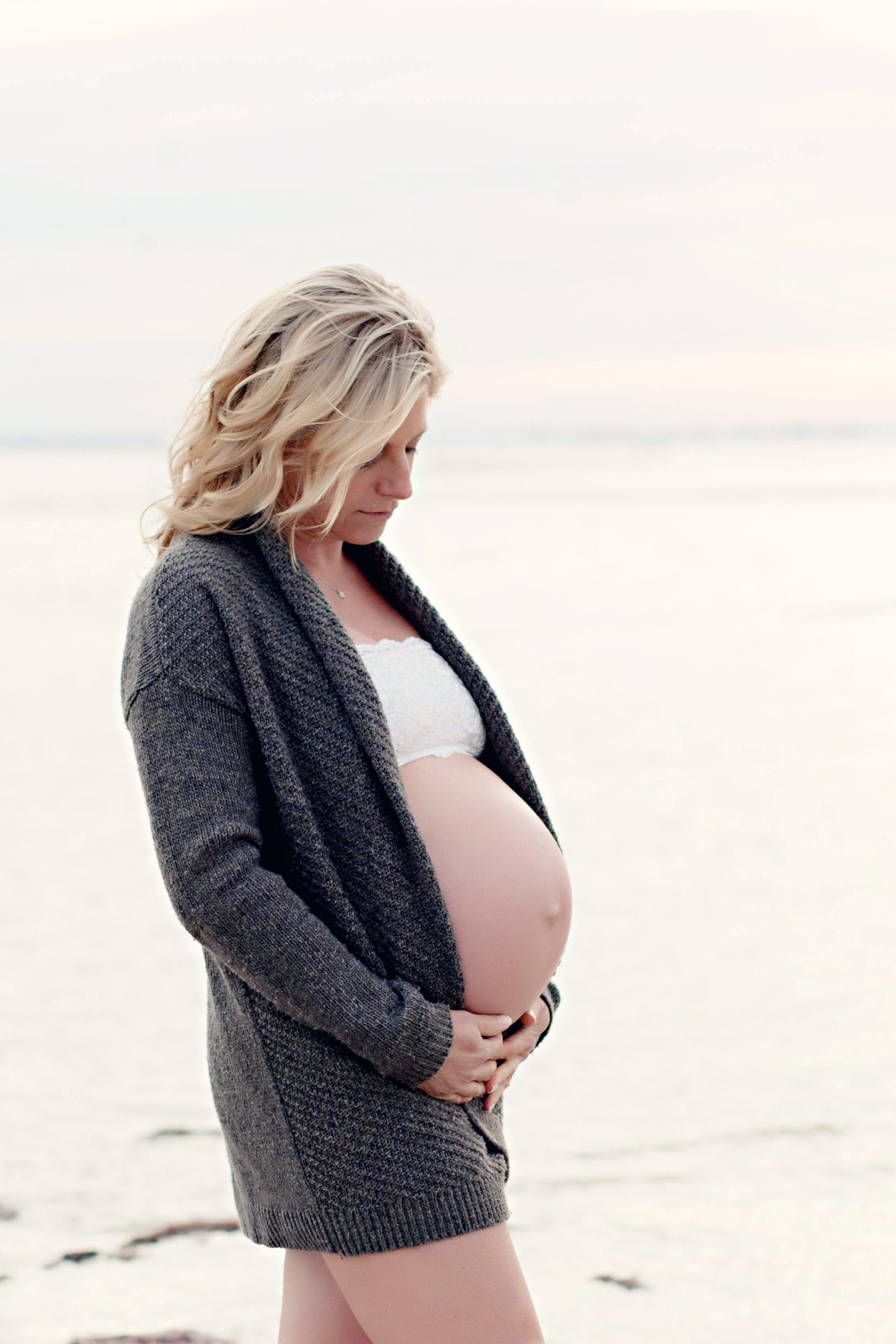 Pregnant blonde woman in gray sweater on a beach, cradling her belly.