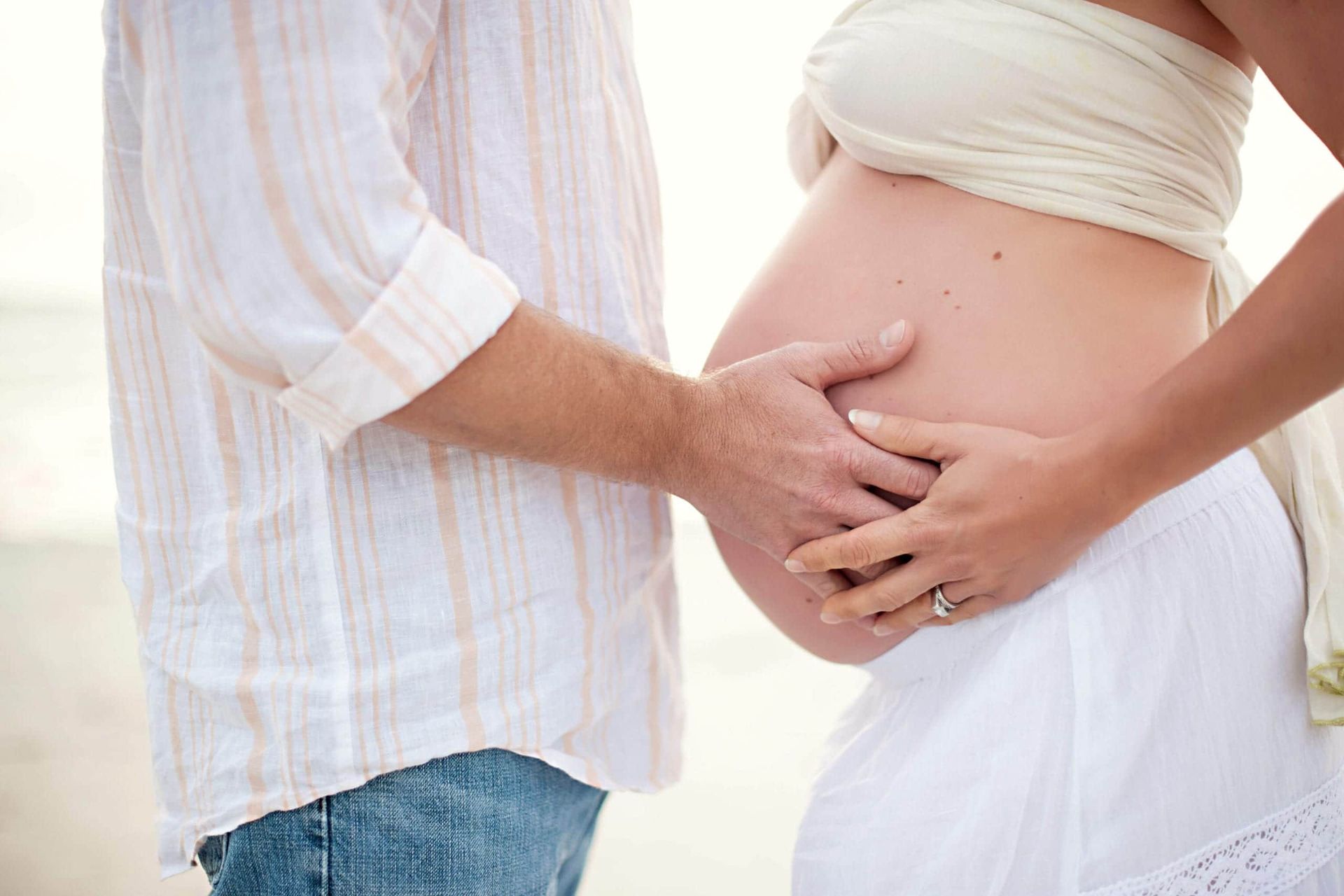 Couple touching pregnant belly at the beach; woman in white, man in light shirt, outdoor daylight.
