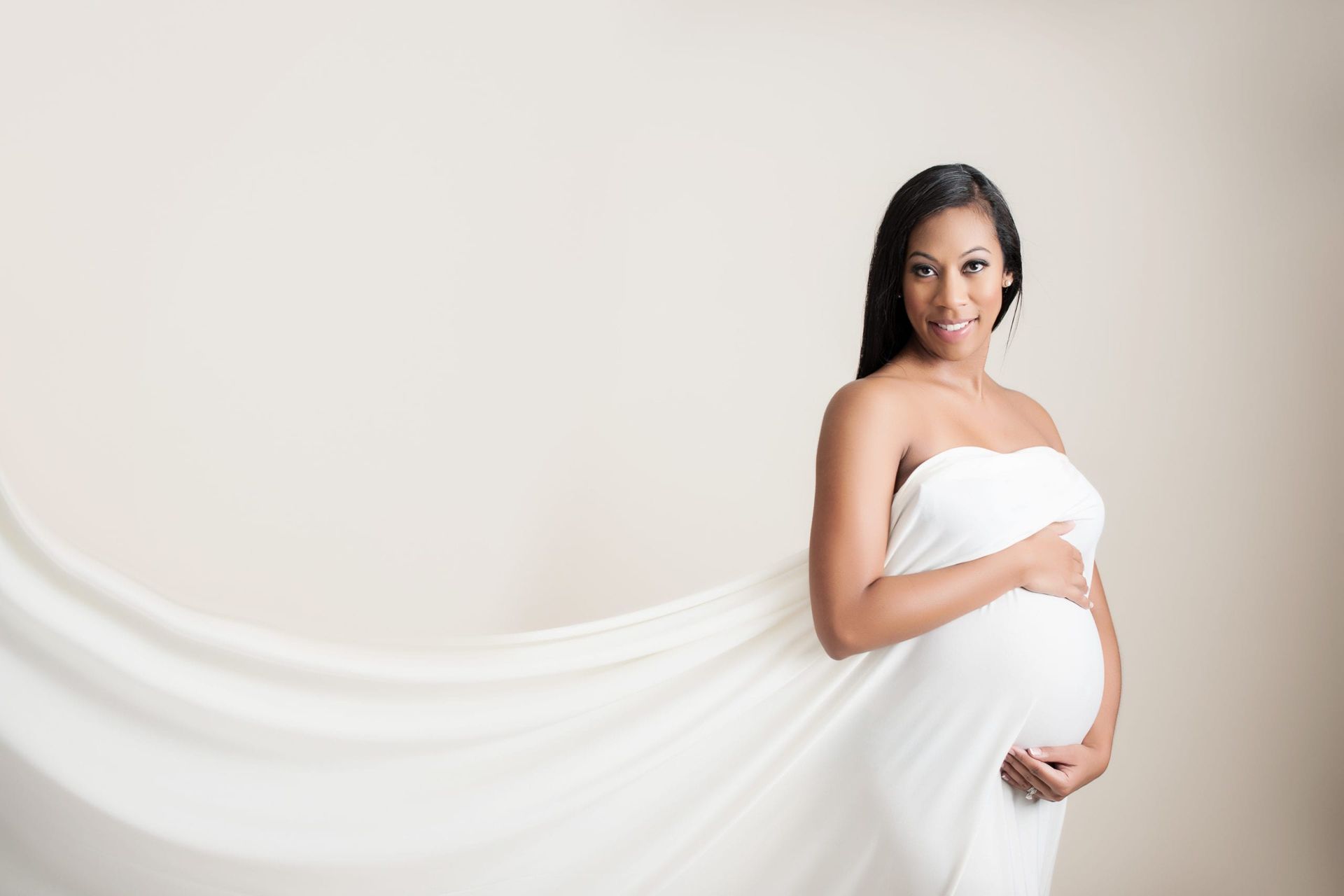 Pregnant woman in white fabric, smiling, holding her belly; cream background.