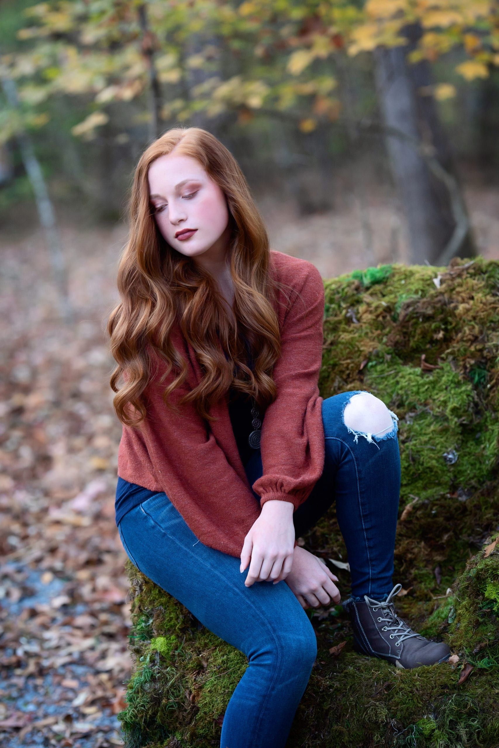 Young woman with red hair, wearing jeans and a rust-colored sweater, sits on a moss-covered rock in a forest.
