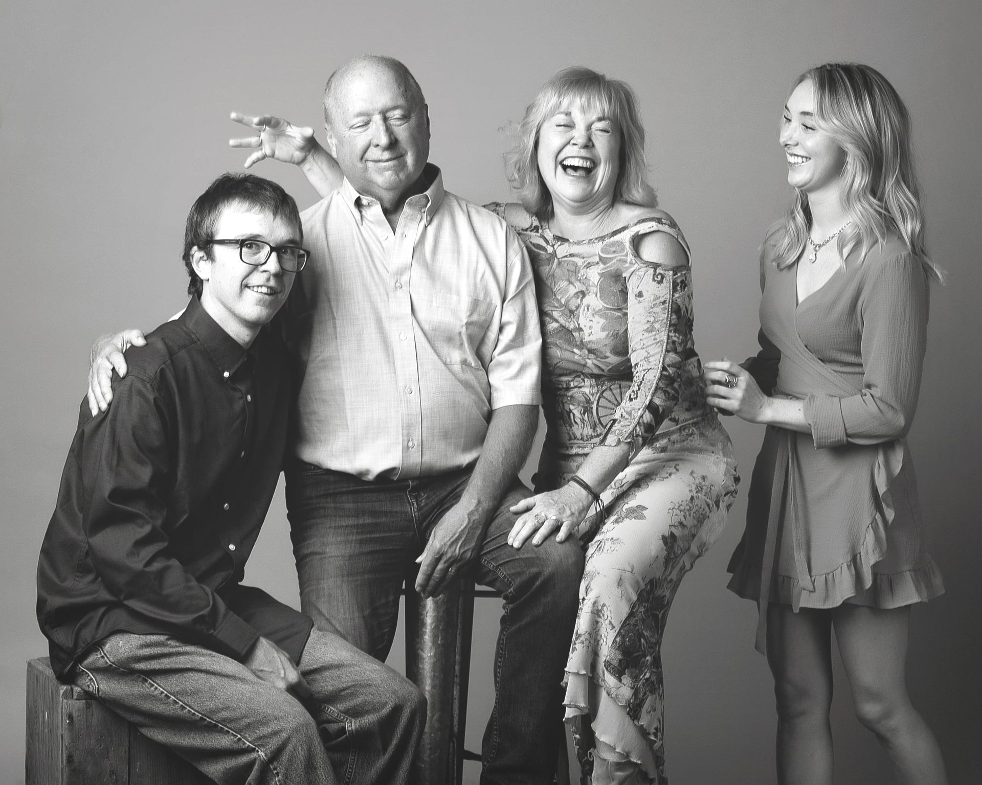 Family of four smiling and laughing; man sits on stool, woman in floral dress, others stand.