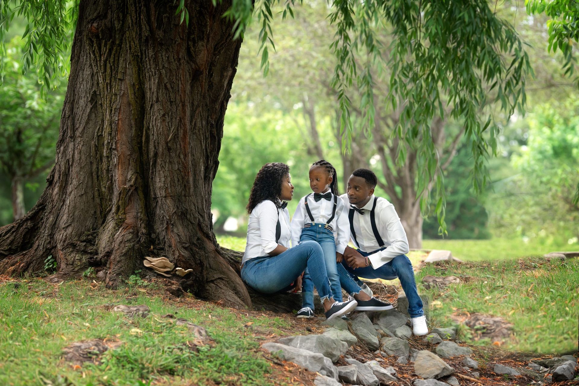 Family of three sits under a large tree, wearing matching outfits, smiling in a park.