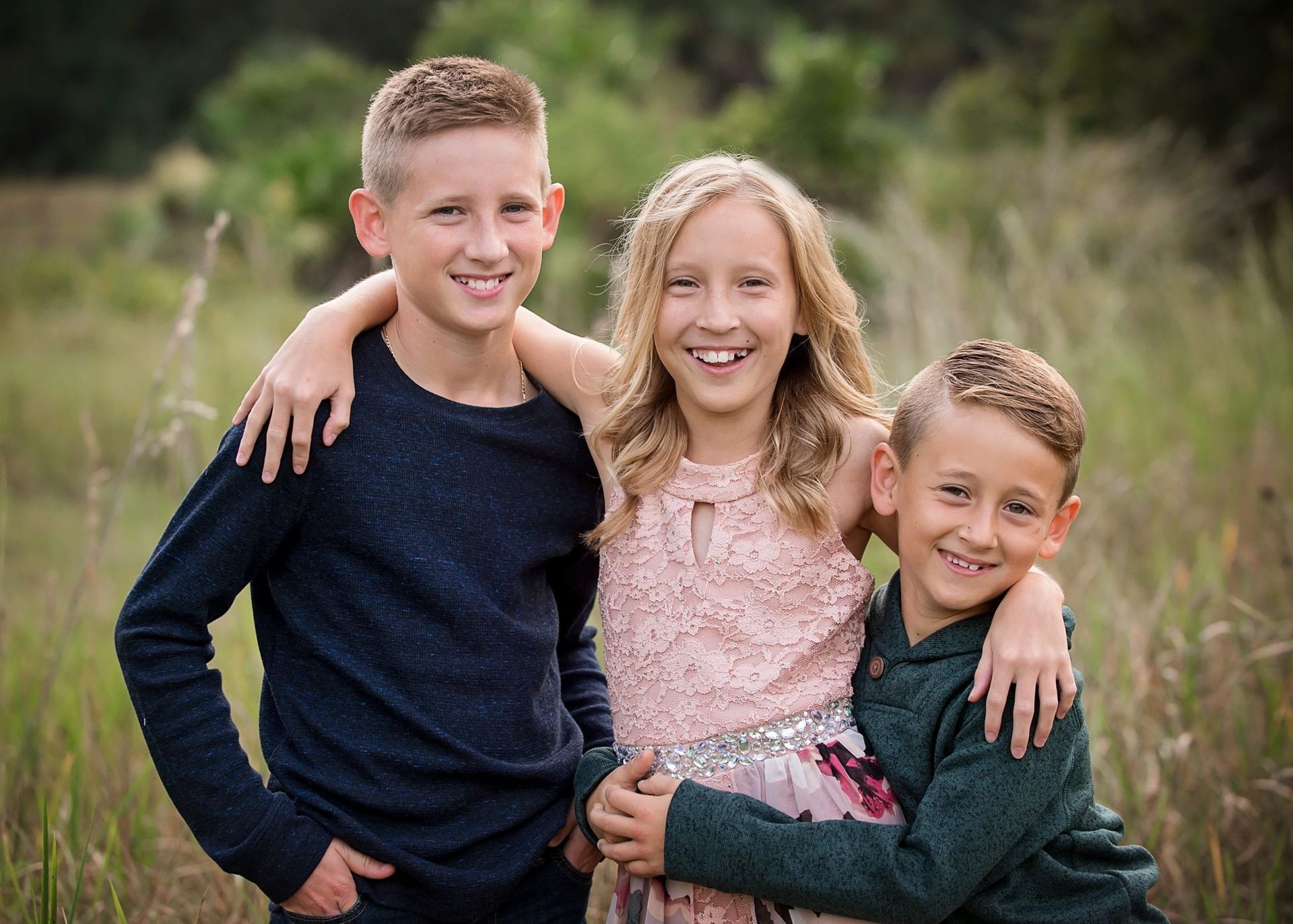 Three smiling children in a field. Two boys and a girl. Boy on left has arm around girl.