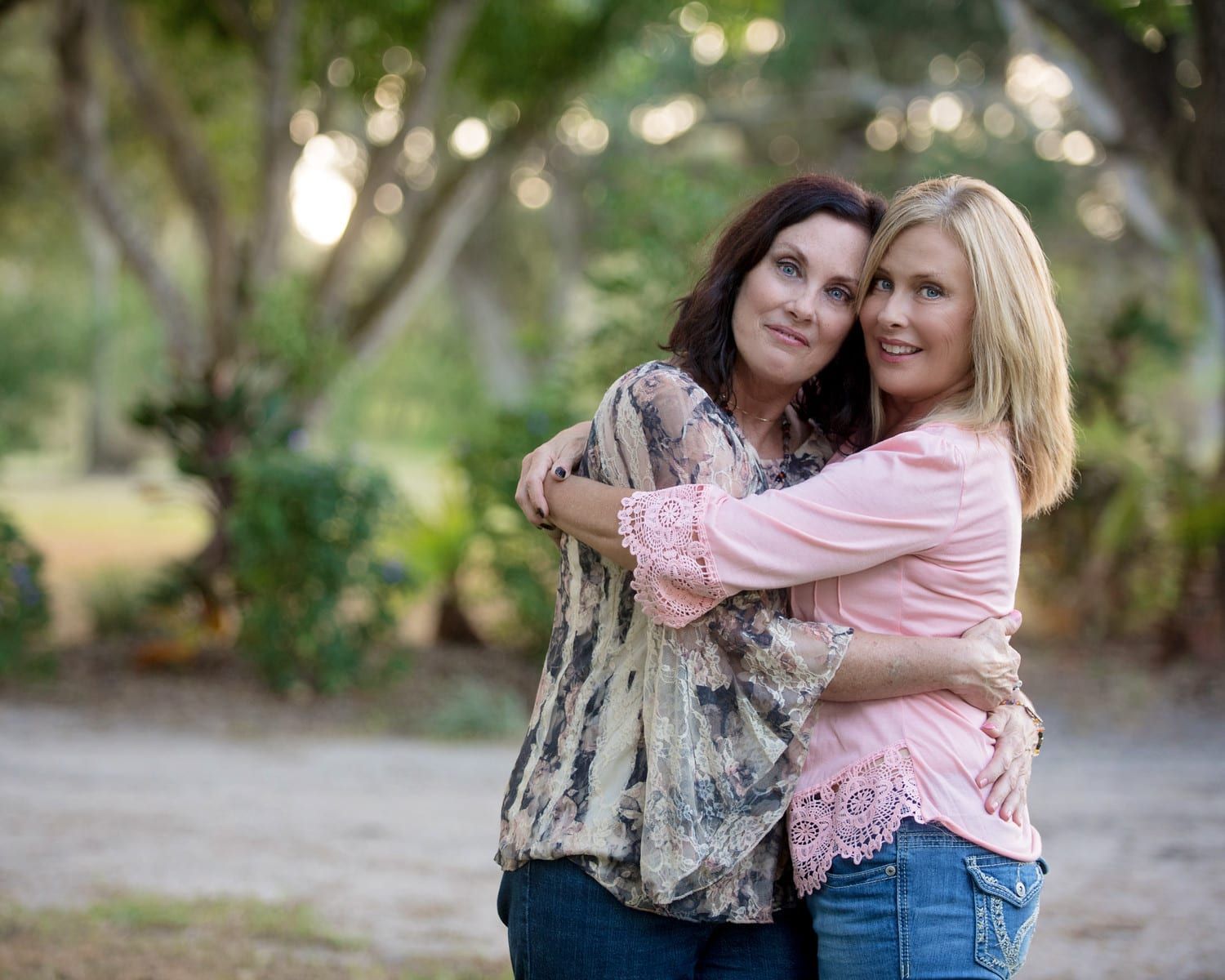 Two women embracing outdoors, smiling. One with dark hair in patterned shirt, the other blonde in pink.