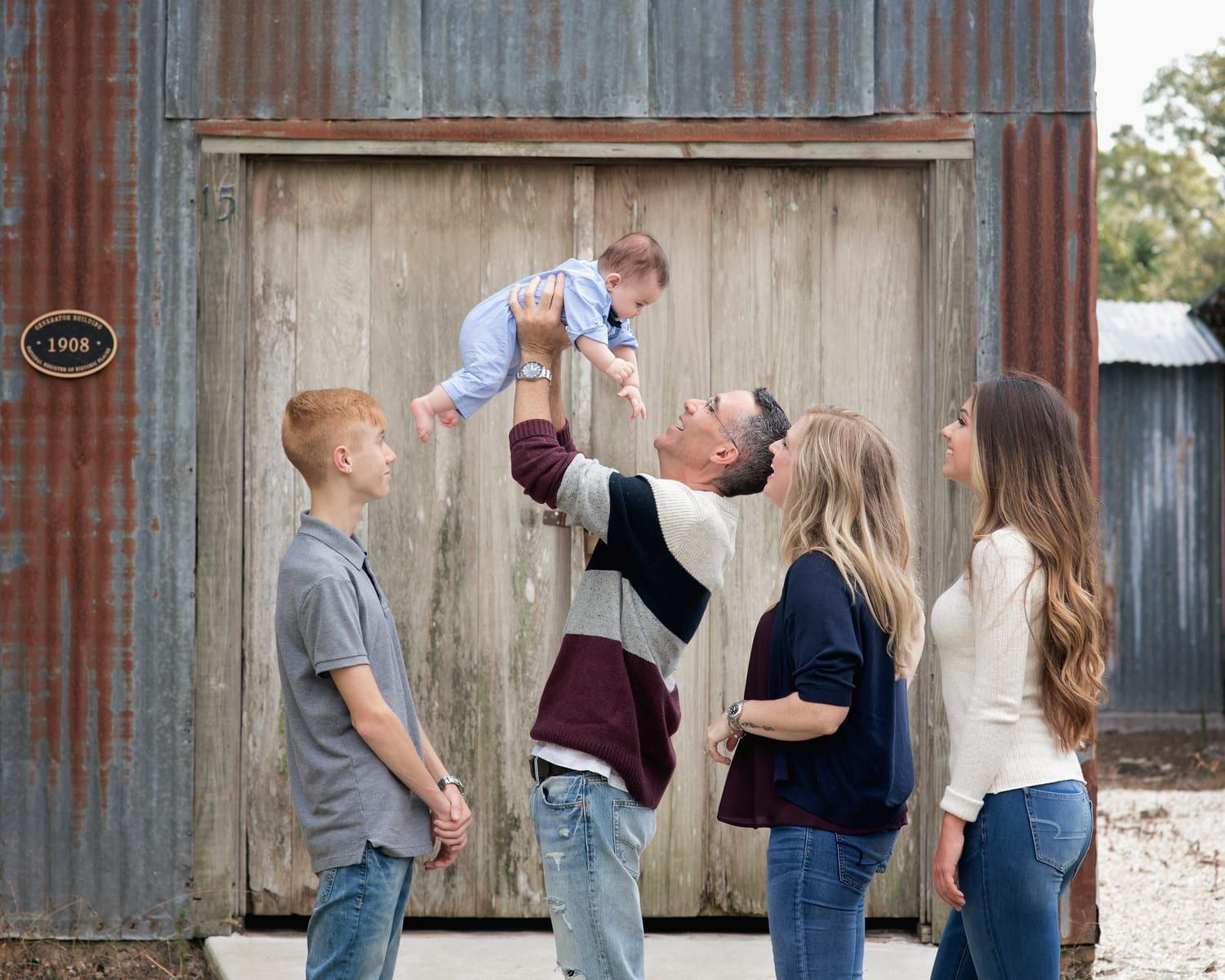 Family of five, father tossing baby up in the air, in front of a weathered wooden building.