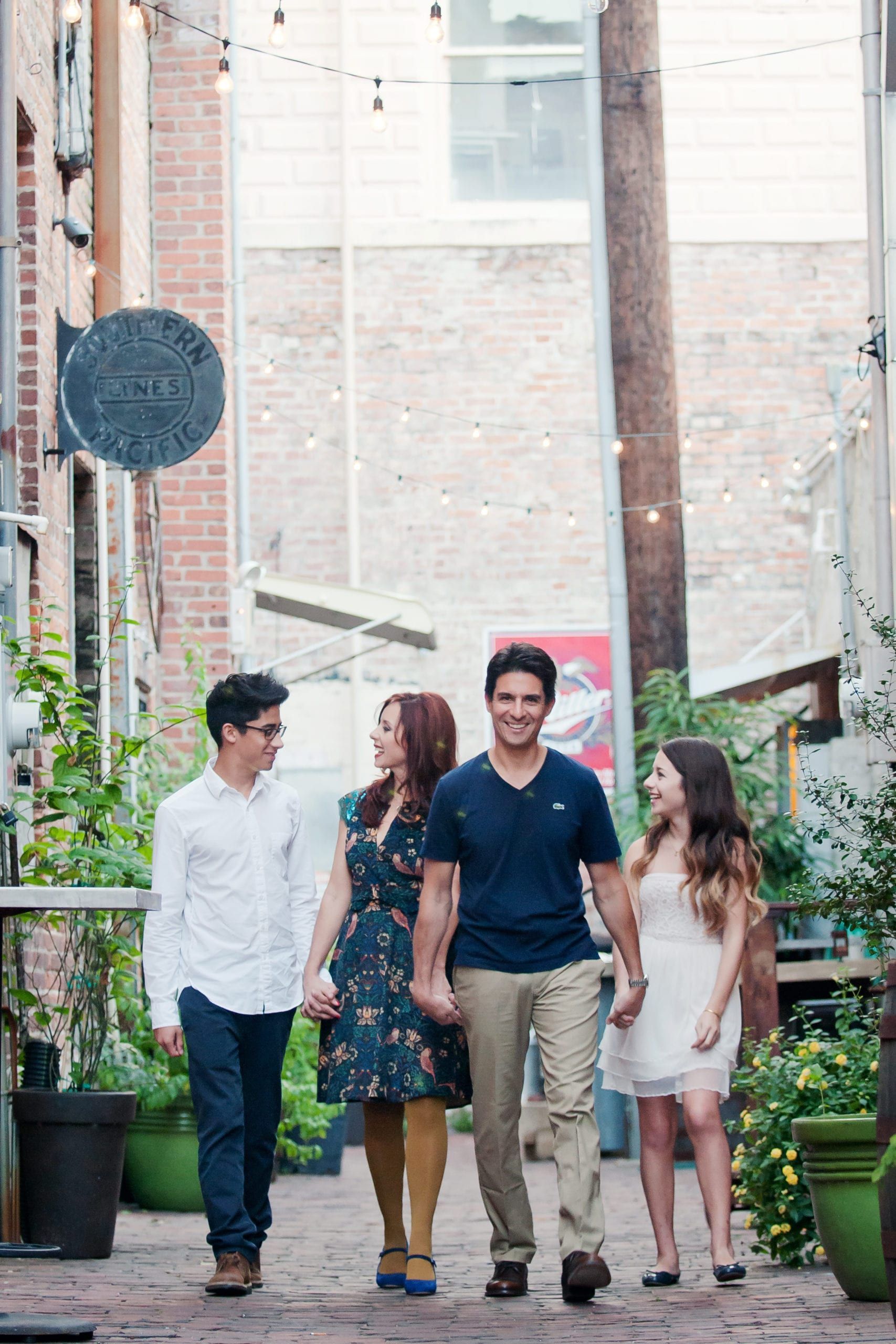 Family of four walking hand-in-hand down an alley, smiling. Brick walls, plants, and string lights visible.