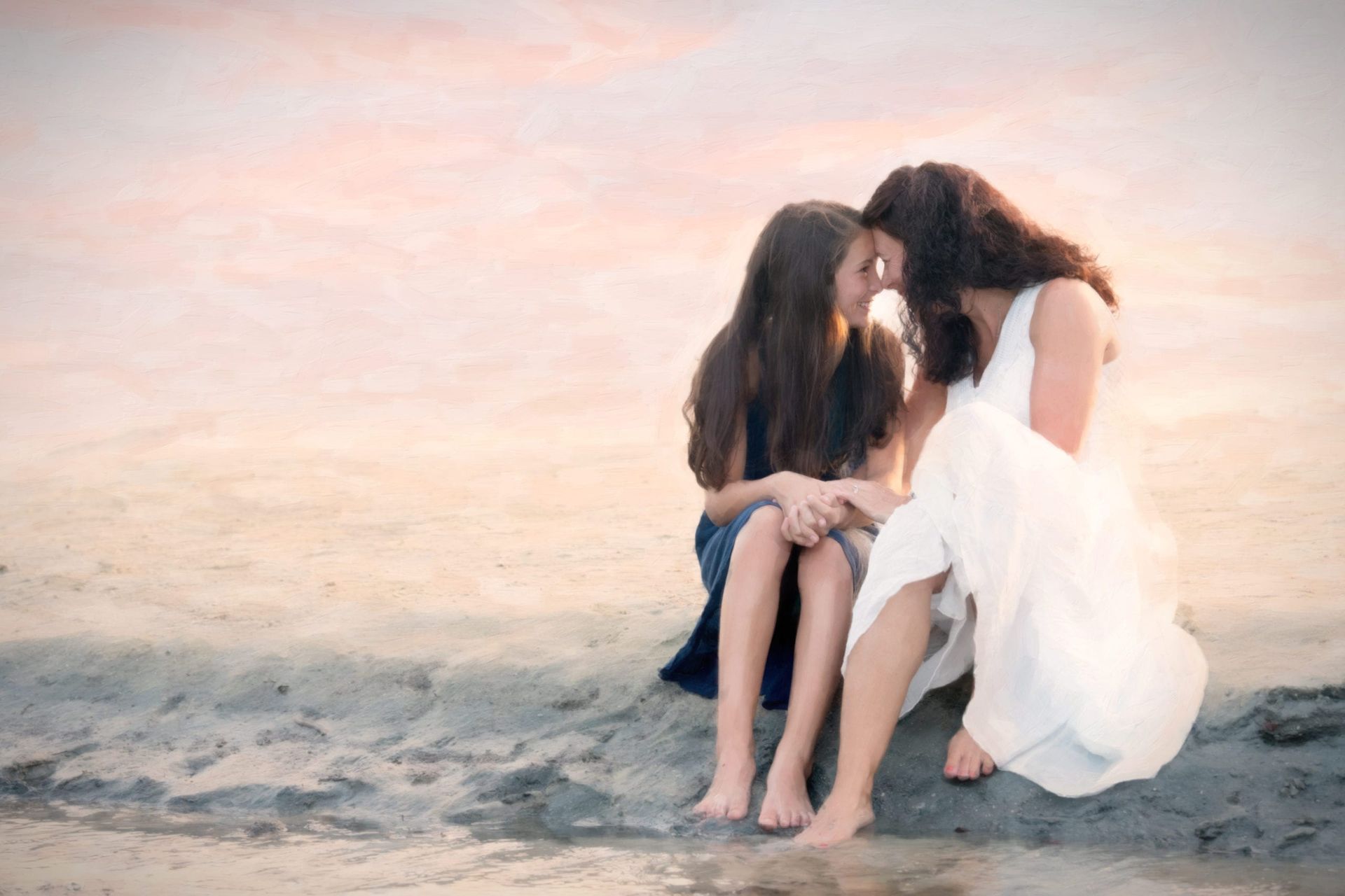 Mother and daughter sit close, faces touching, on a beach at sunset.