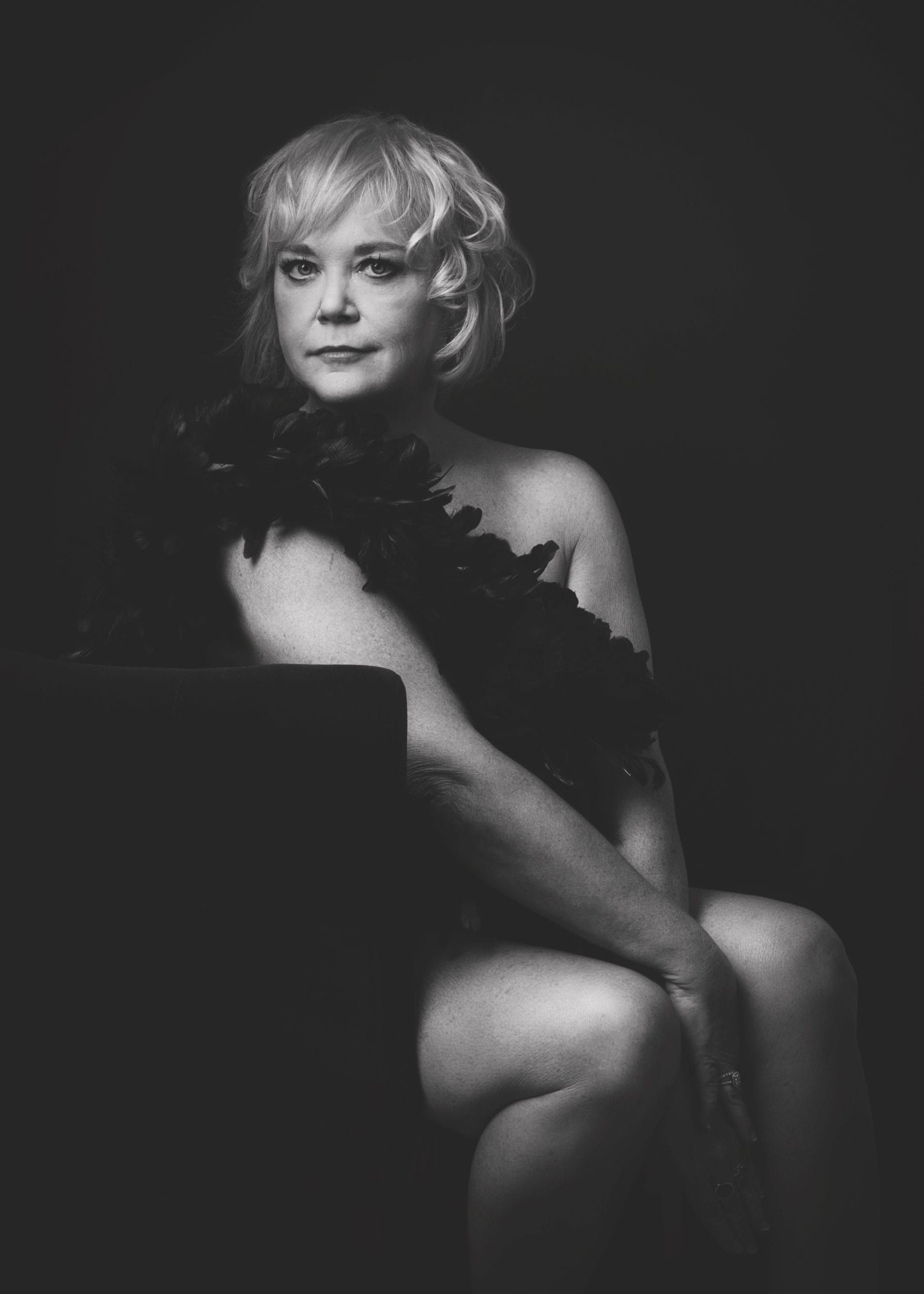 Woman with blonde hair, sitting, nude with a feather boa. Black and white photo.