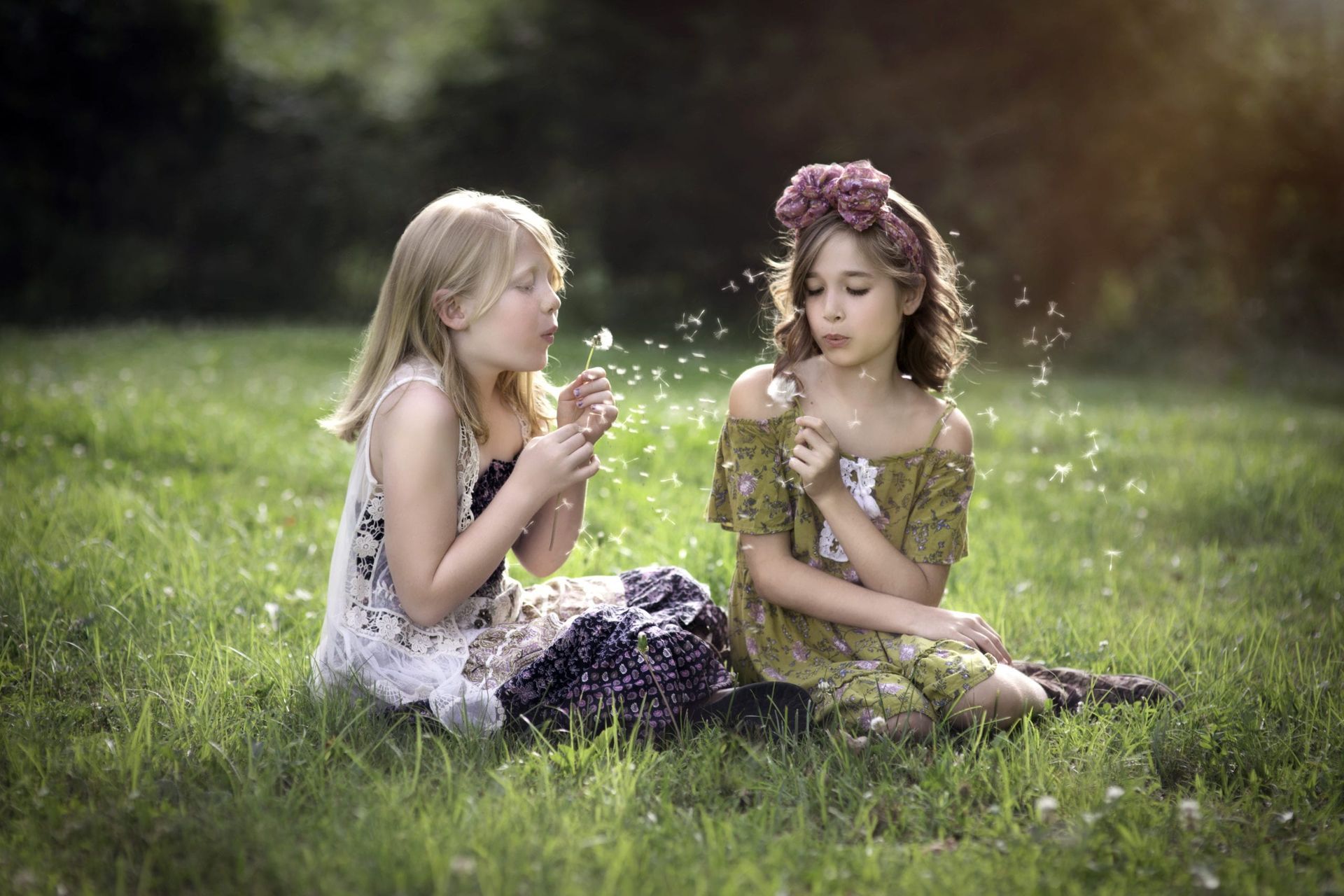 Two young girls blowing dandelion seeds in a grassy field on a sunny day.