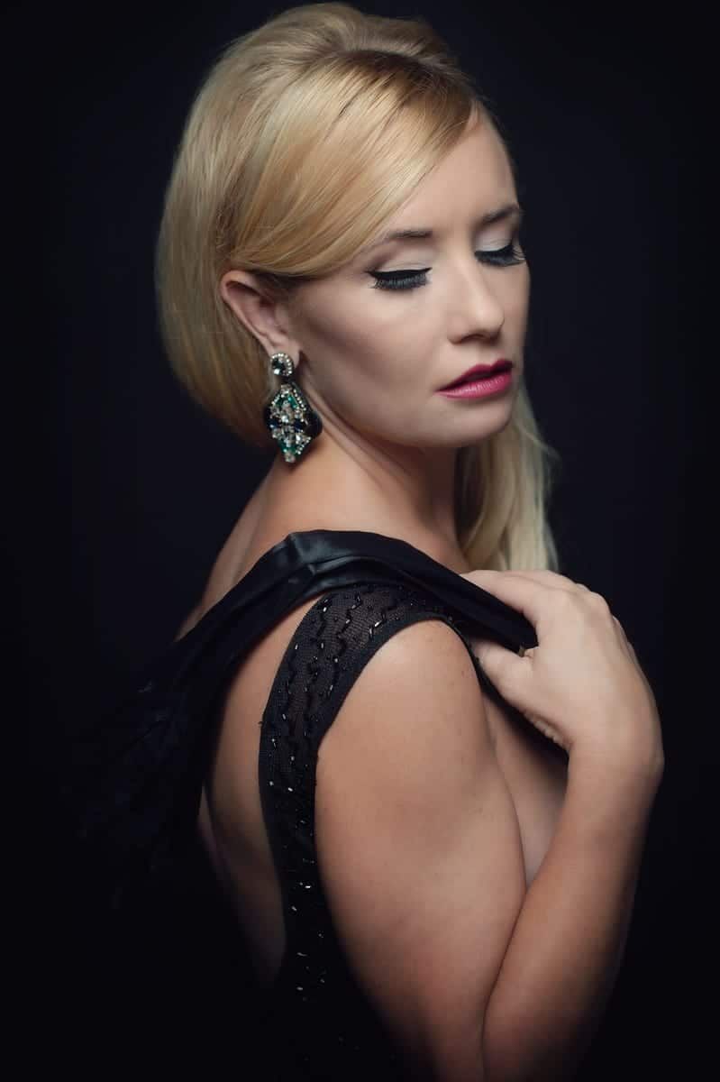 Blonde woman in a black dress, looking down. She has on earrings and is touching her shoulder.