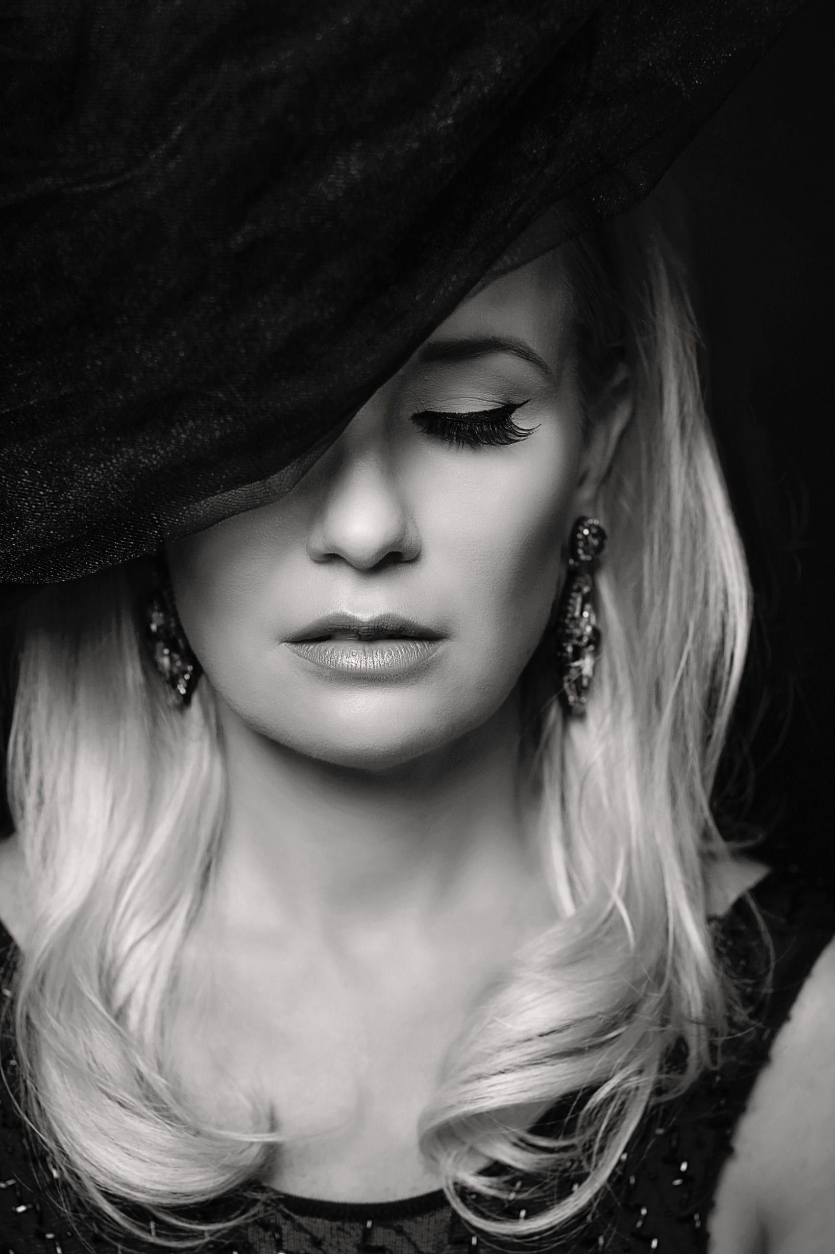Woman with blonde hair, wearing a black hat with veil, earrings. Black and white portrait, contemplative expression.