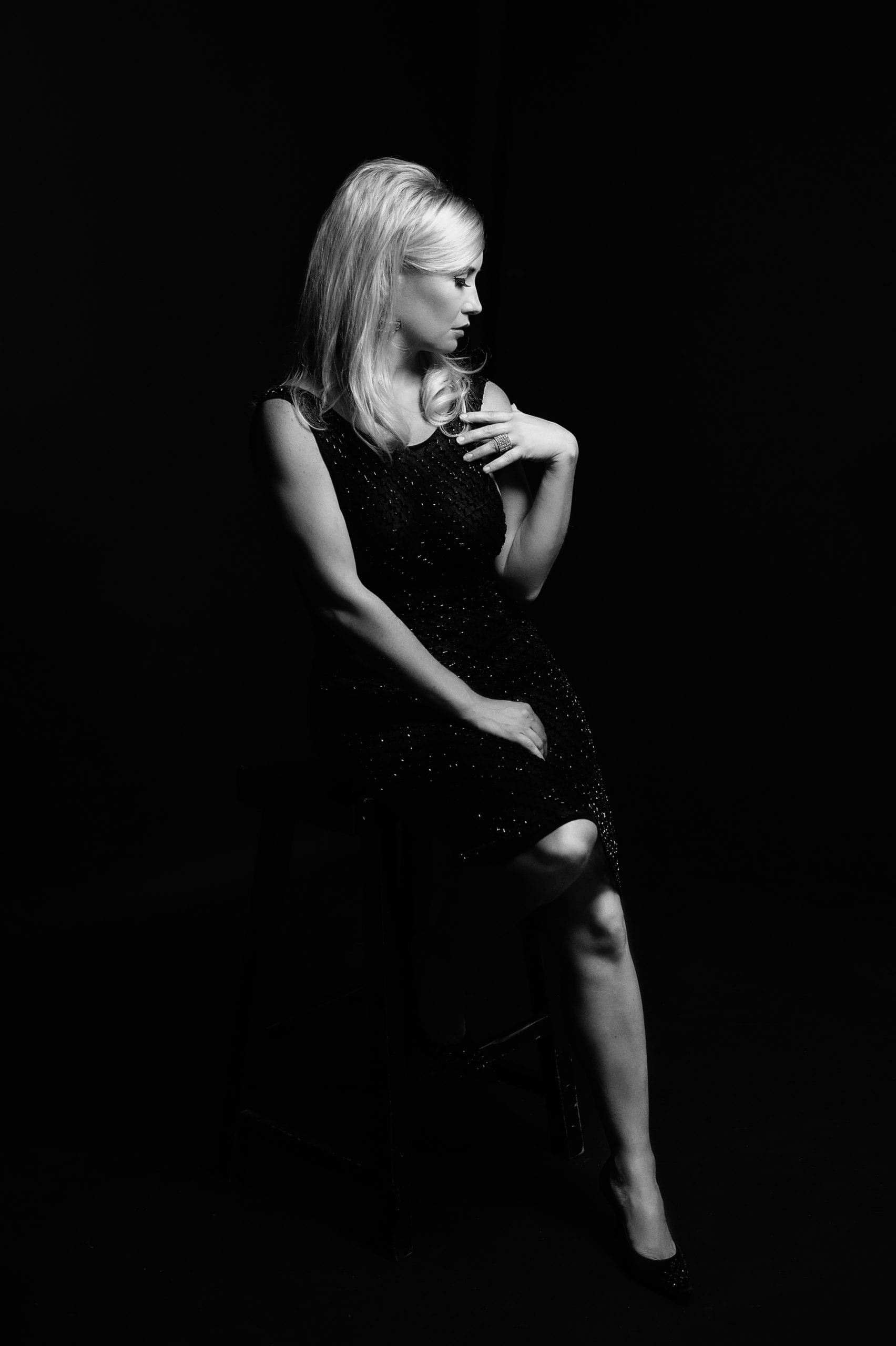 Blonde woman in a sparkling black dress sits, angled away, hand on chest. Dark background.