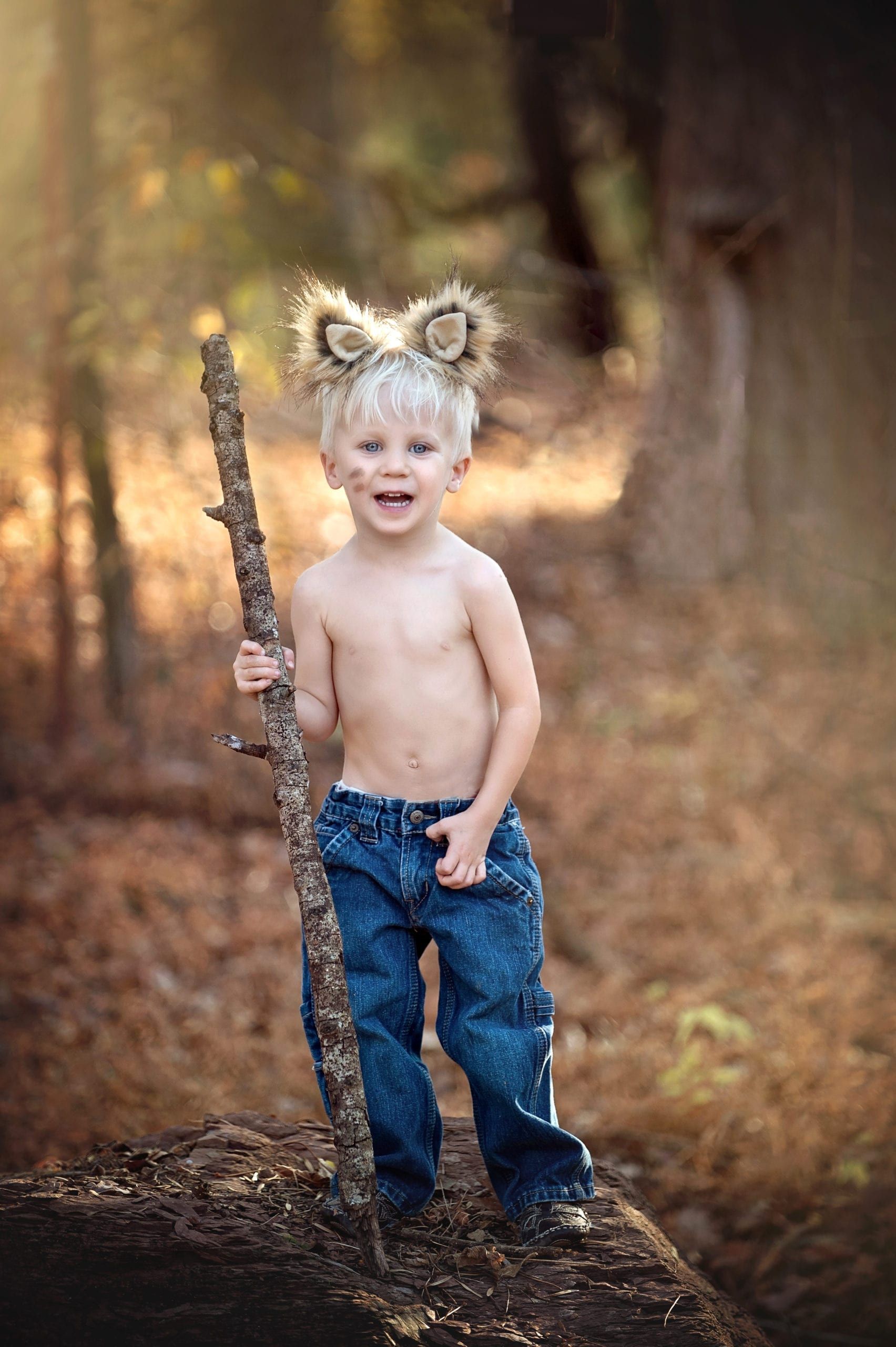 Blond child with furry ears, holding a stick, smiling in a forest setting, wearing jeans and shirtless.