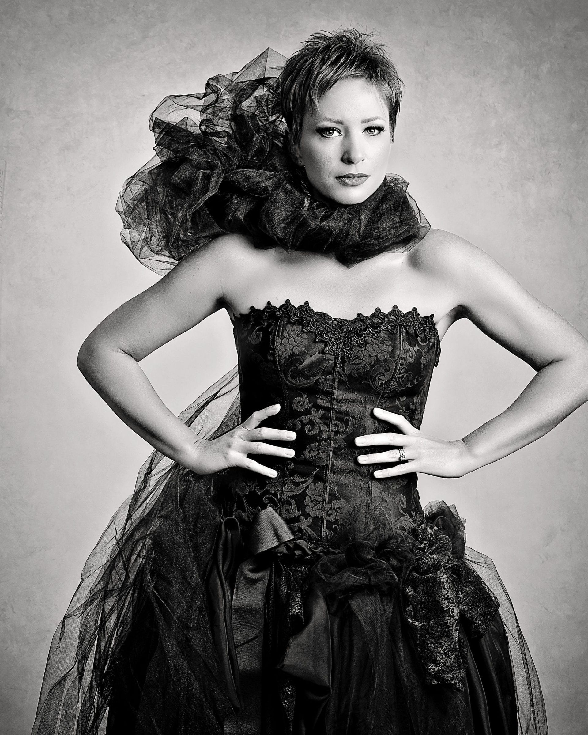 Woman in elaborate black gown with arms on hips, looking forward.