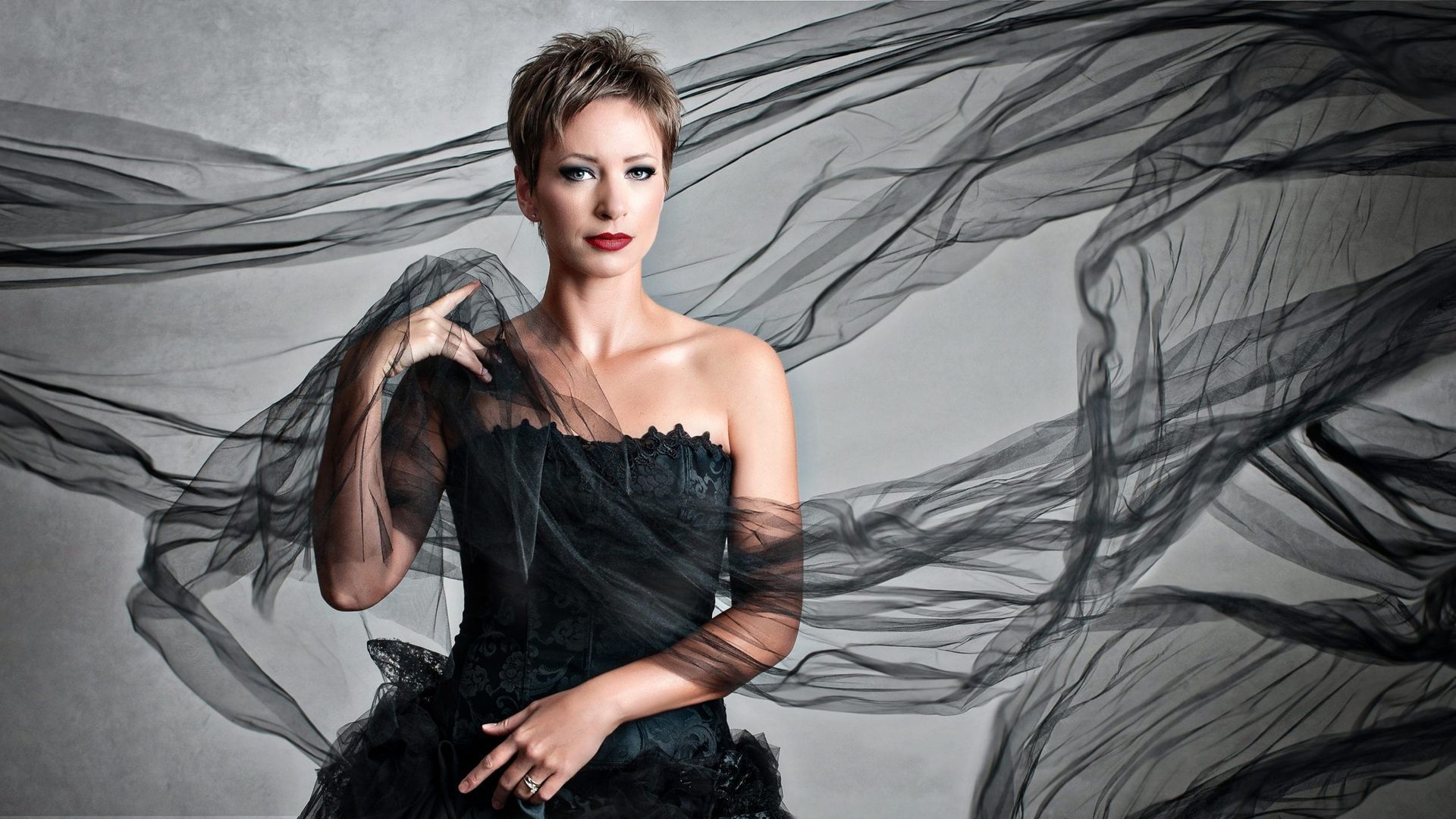 Woman in black dress, short hair, looking forward, with flowing dark fabric against a grey background.