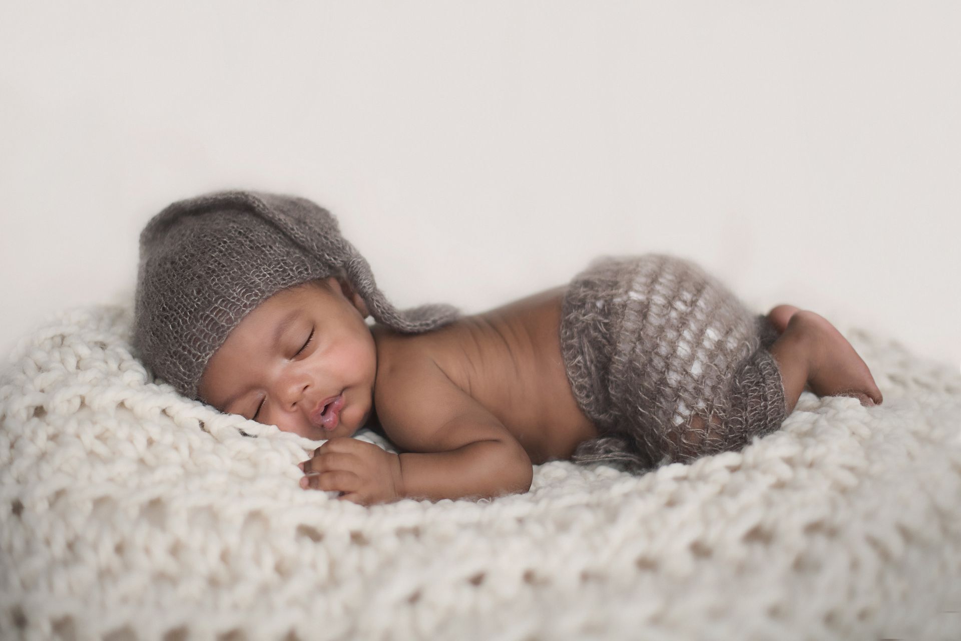Newborn baby sleeping on a white knitted blanket, wearing a gray hat and shorts.