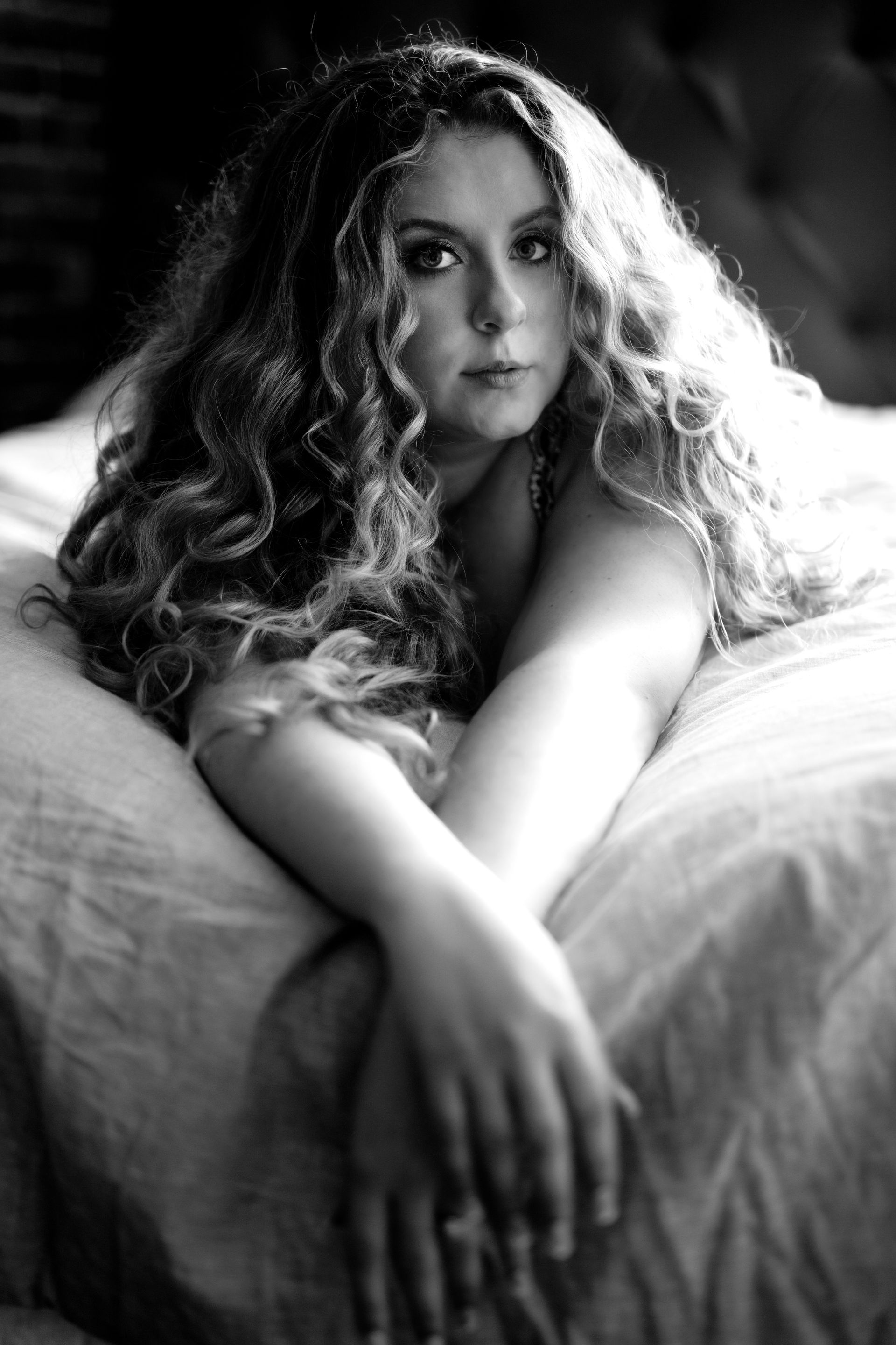 Woman with long curly hair, lying on a bed, arms crossed, looking at the viewer. Black and white.