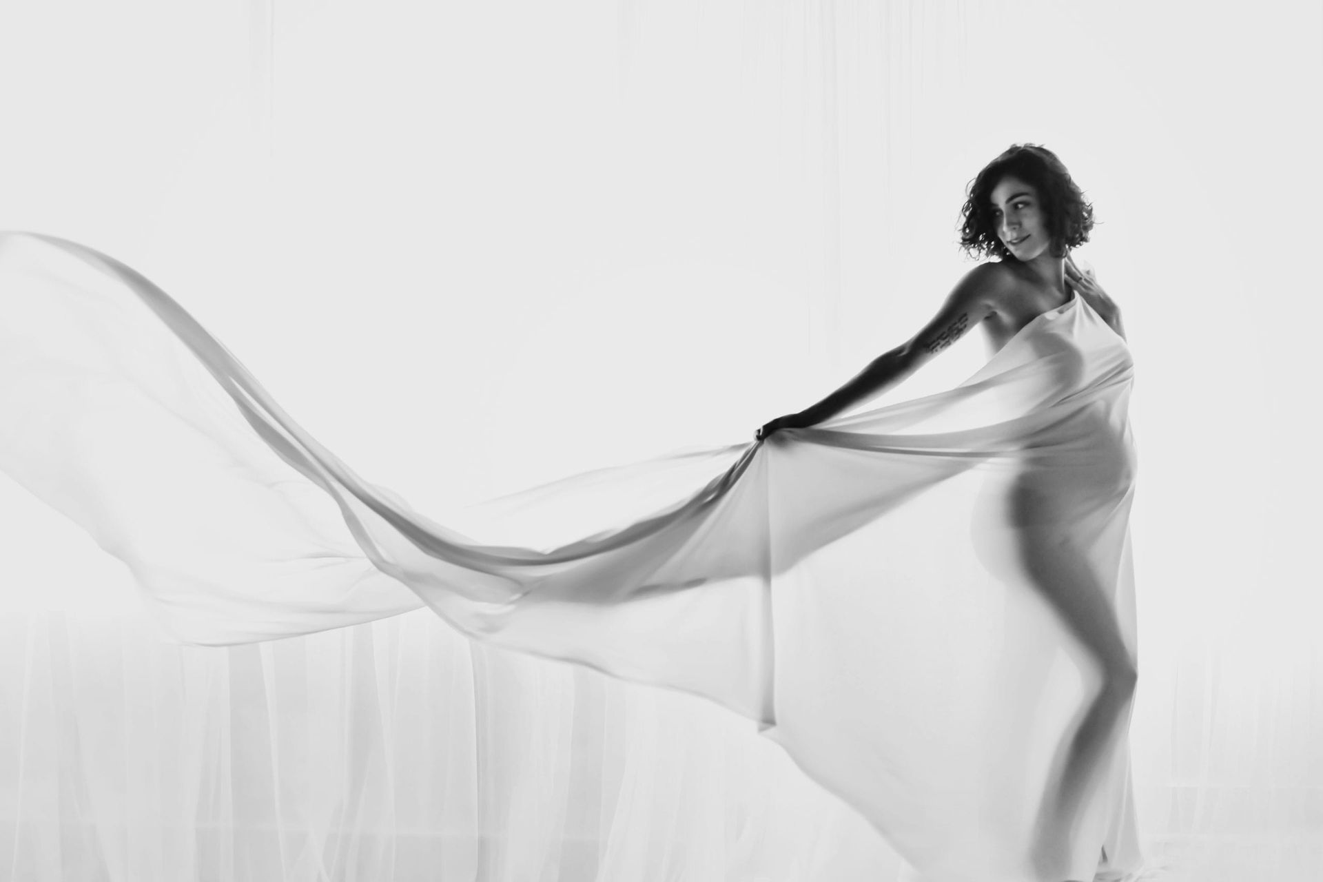 Pregnant woman in flowing sheer fabric, silhouetted against a bright white background, looking to the side.