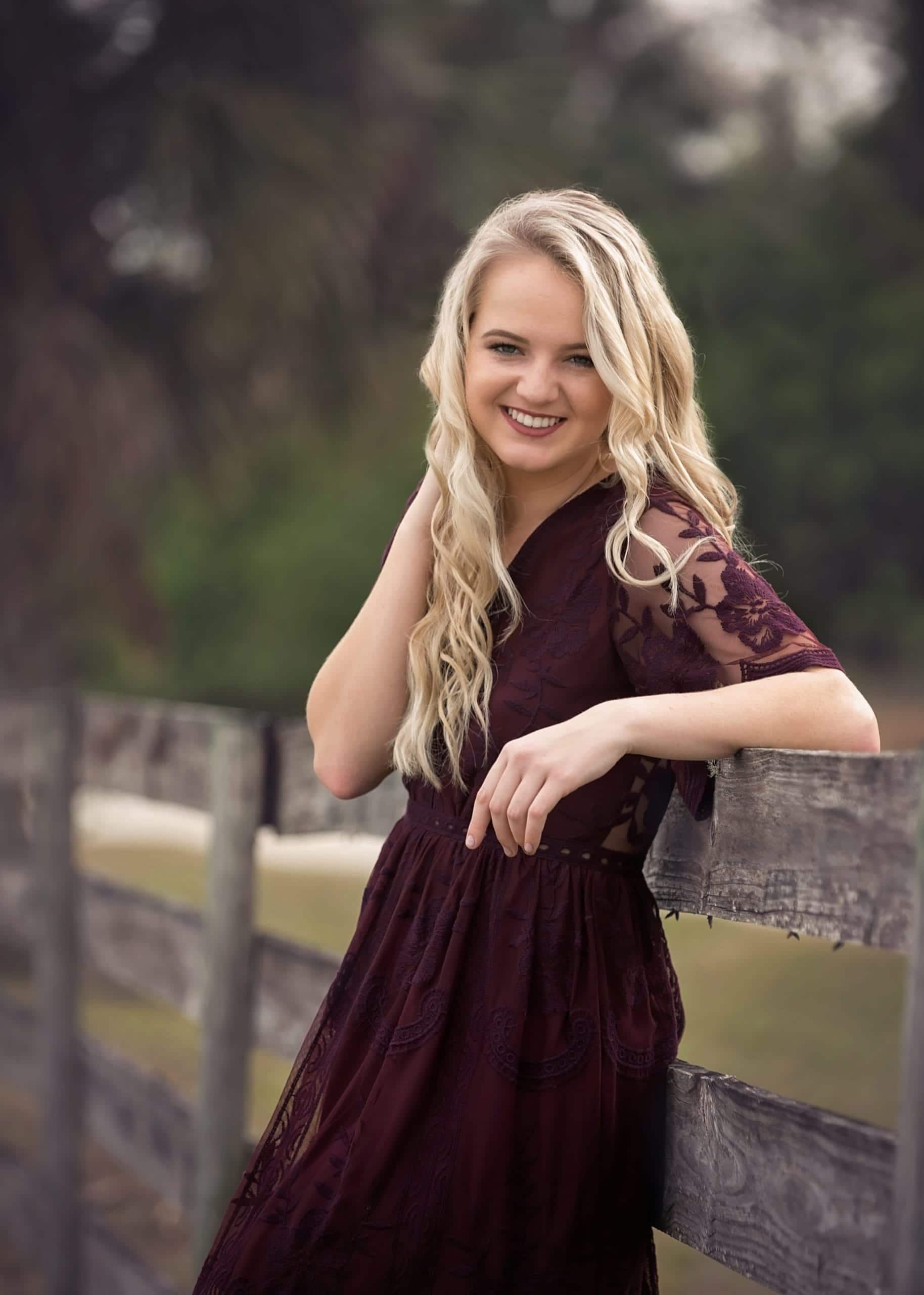 Blonde woman in maroon lace dress leans on a wooden fence, smiling outdoors.