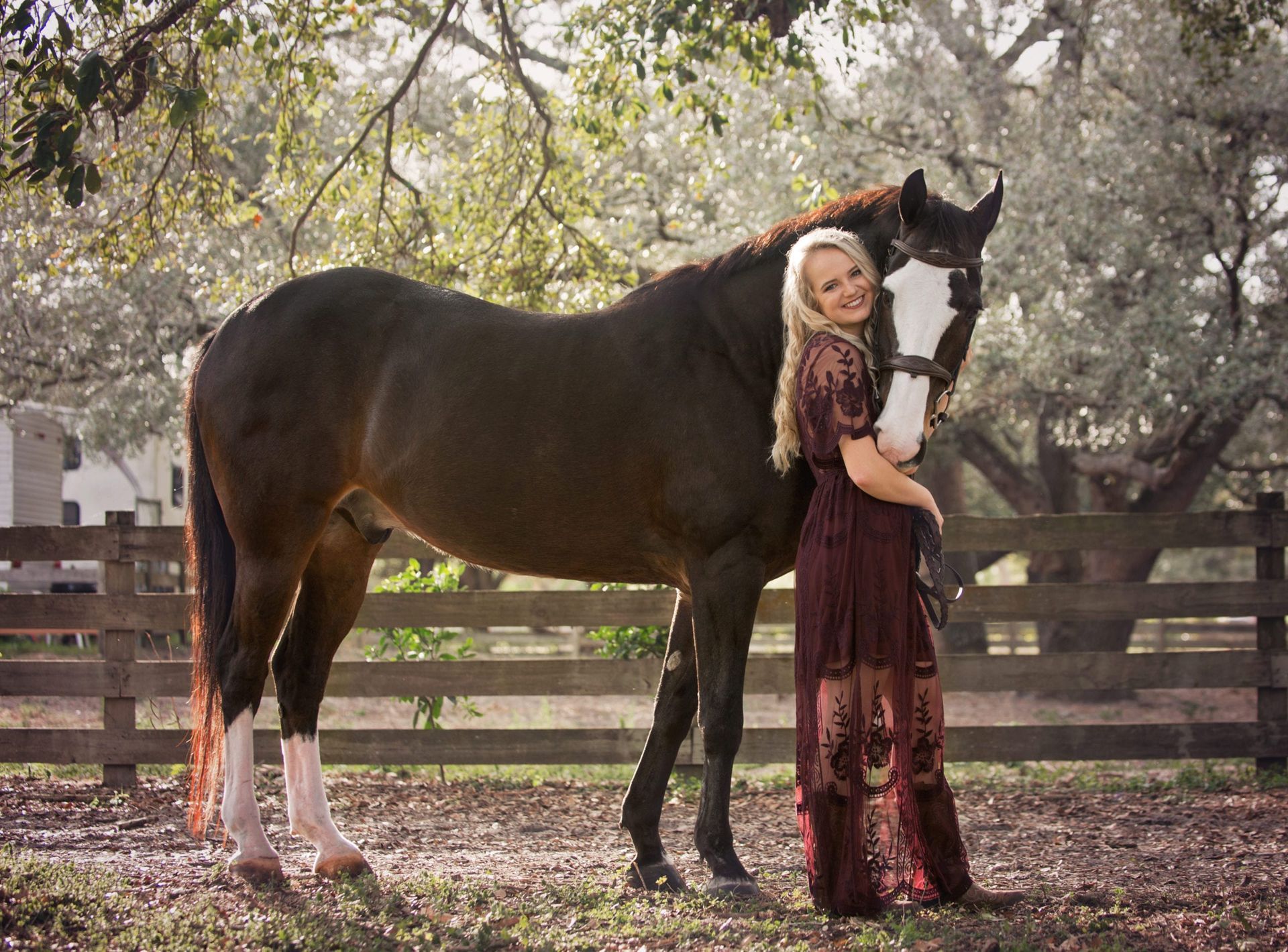 Woman in burgundy dress smiles, hugging brown horse with white face and legs, by a wooden fence.