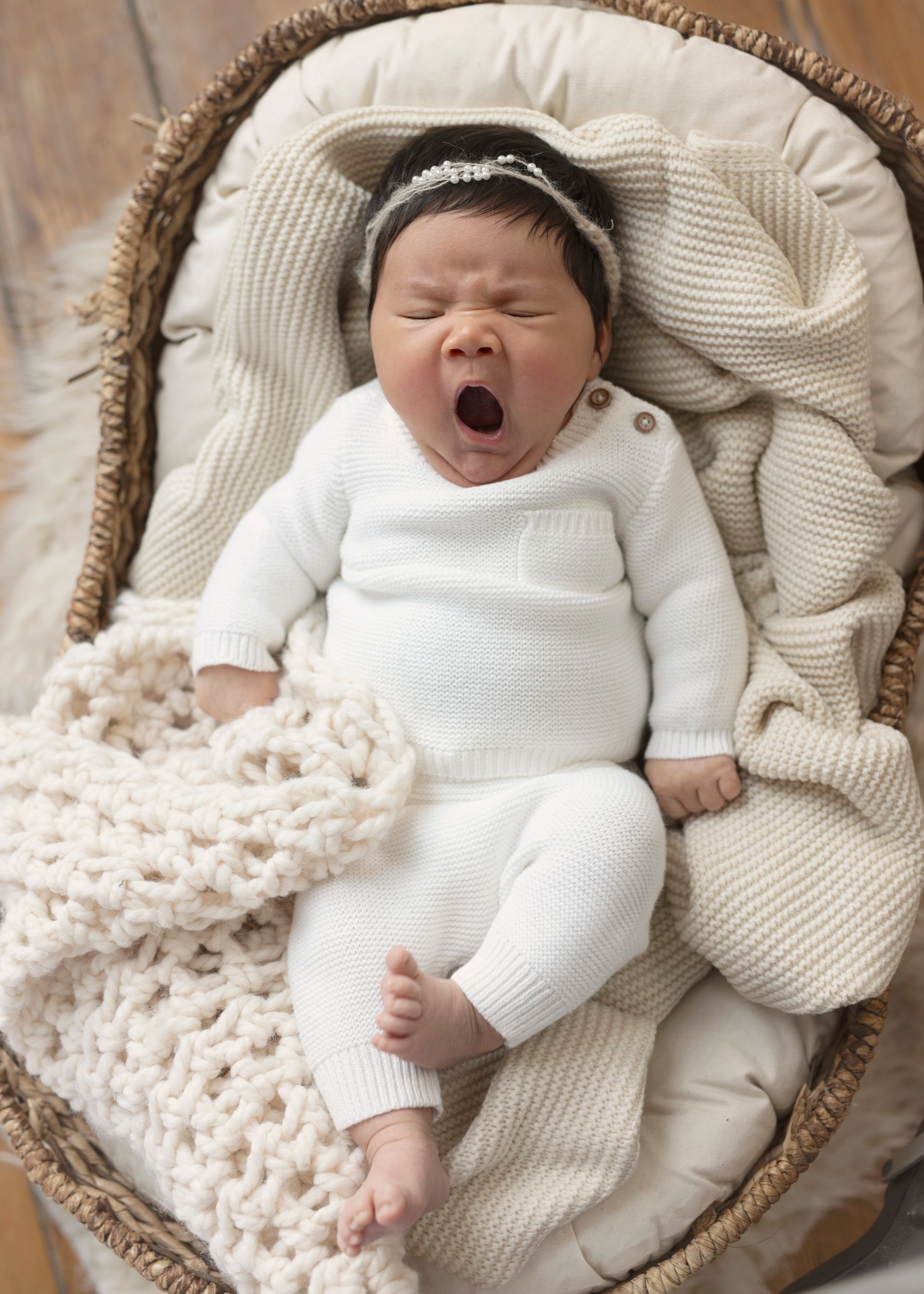 Newborn baby yawning, dressed in white knitwear, in a woven basket with soft blankets.