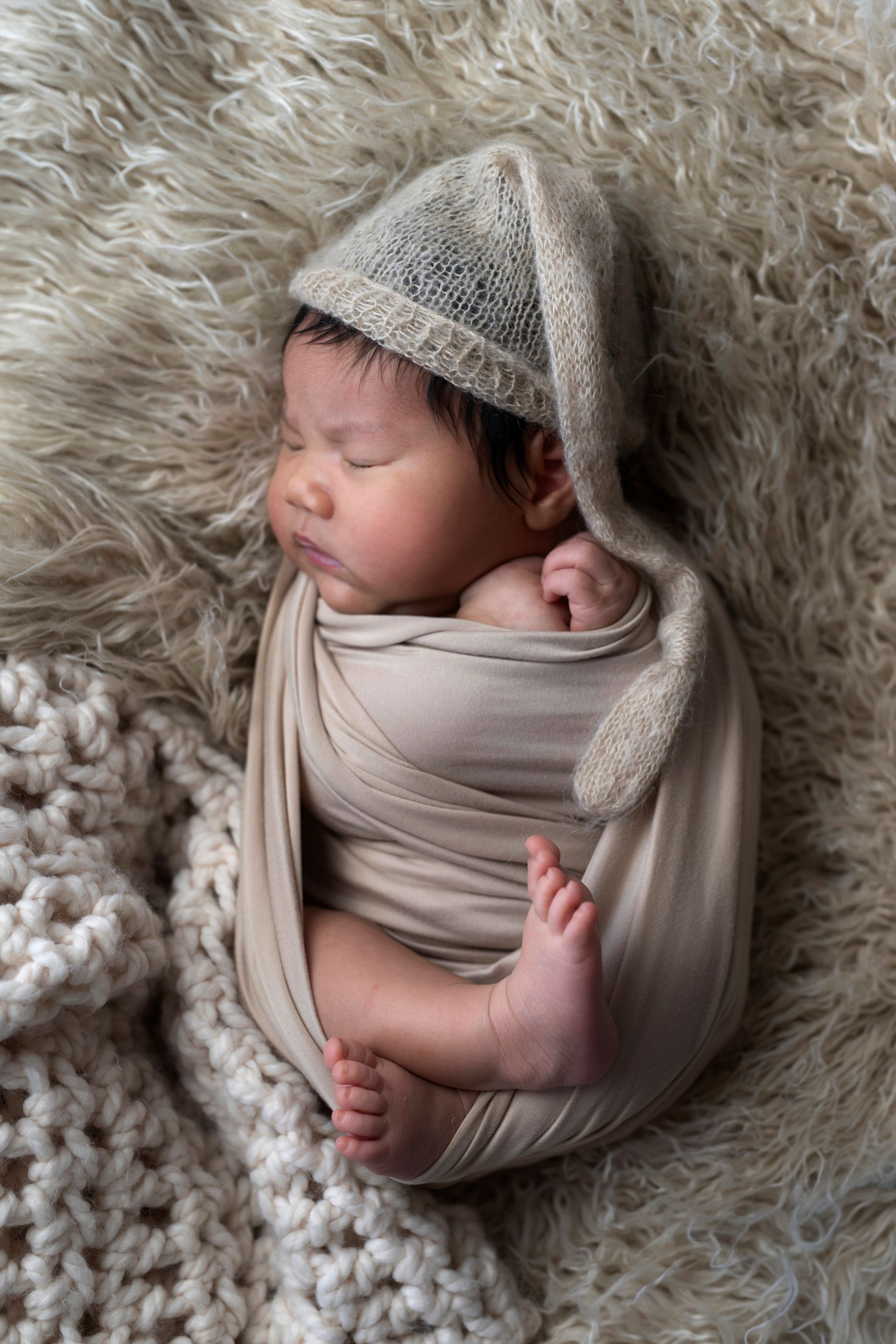 Newborn baby wrapped in tan swaddle, wearing a knitted hat, sleeping on fur blanket.