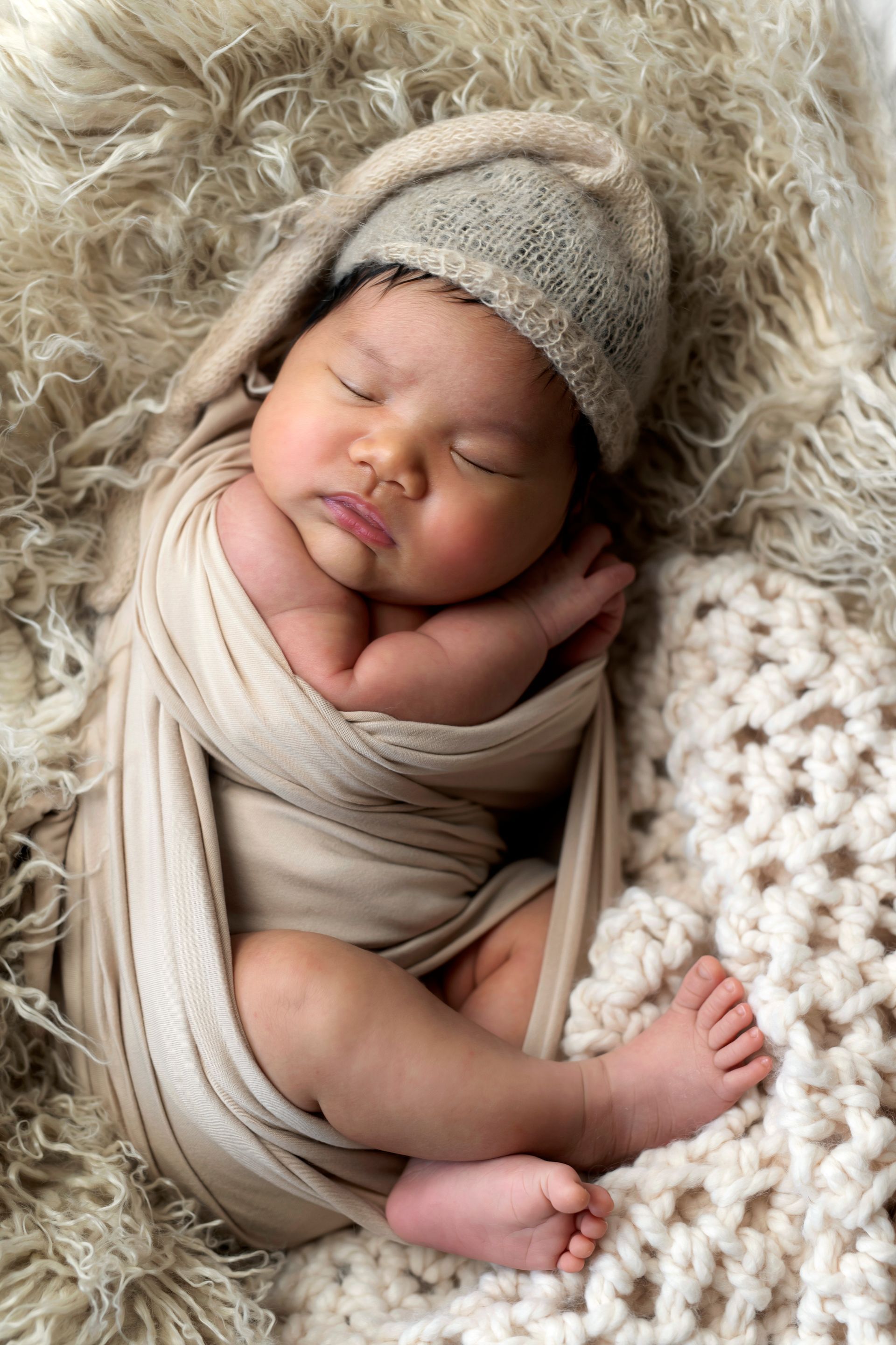 Sleeping newborn swaddled in beige, wearing a knit cap, resting on a textured cream blanket.