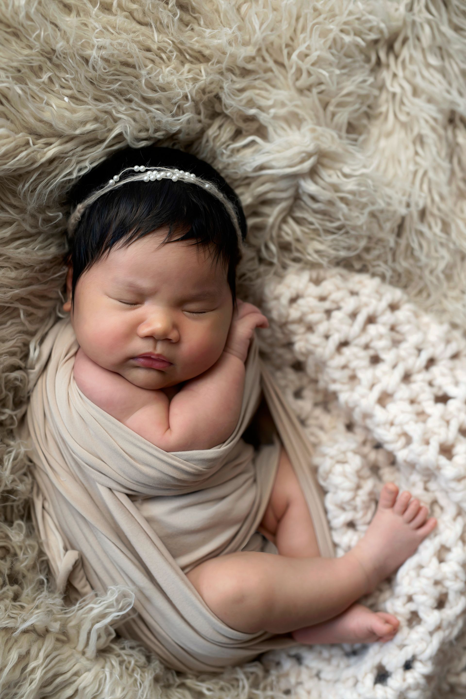 Newborn baby wrapped in beige fabric, sleeping on a fluffy, light brown blanket.