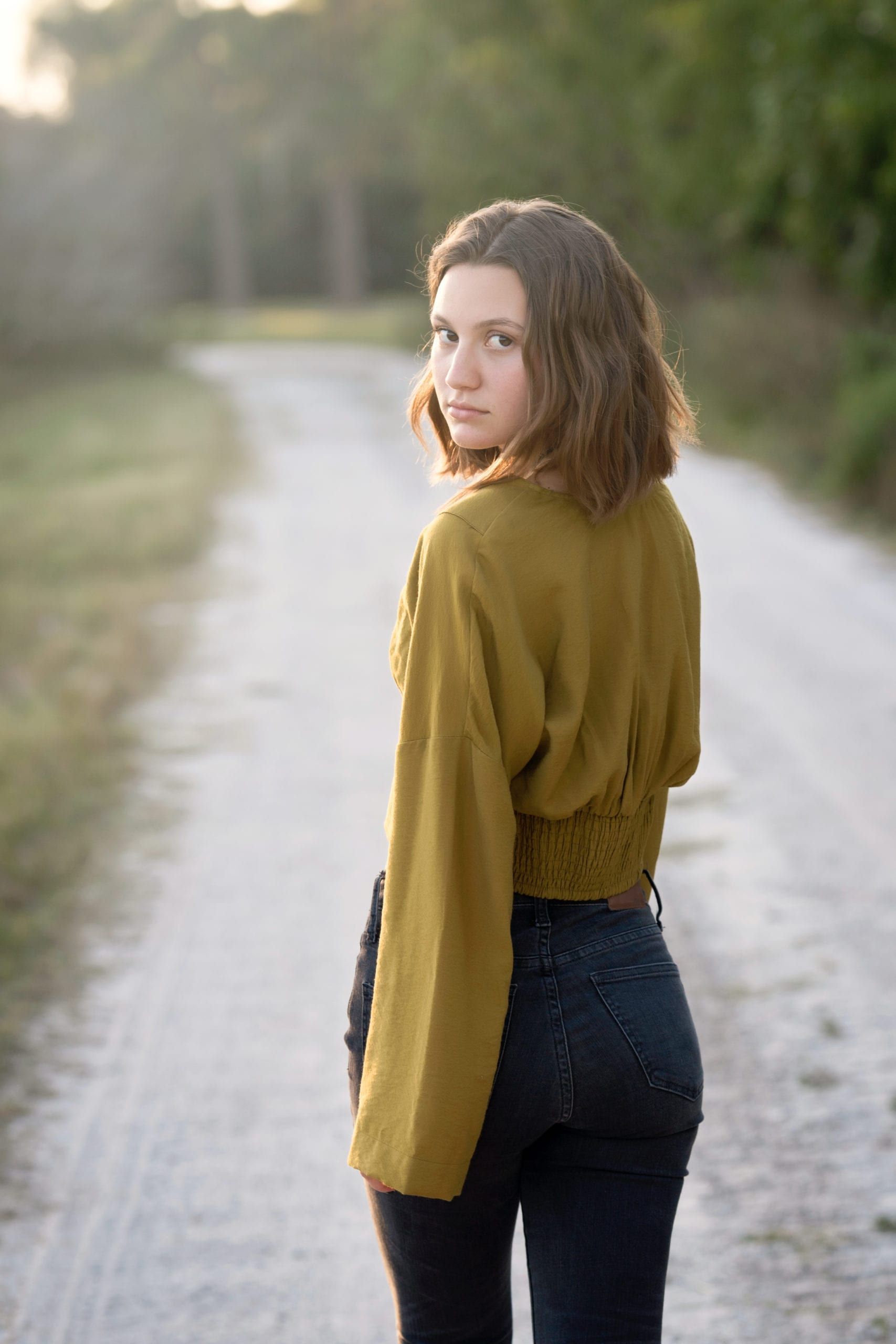 Woman with short brown hair looks over her shoulder on a dirt road. She wears a mustard sweater and dark jeans.